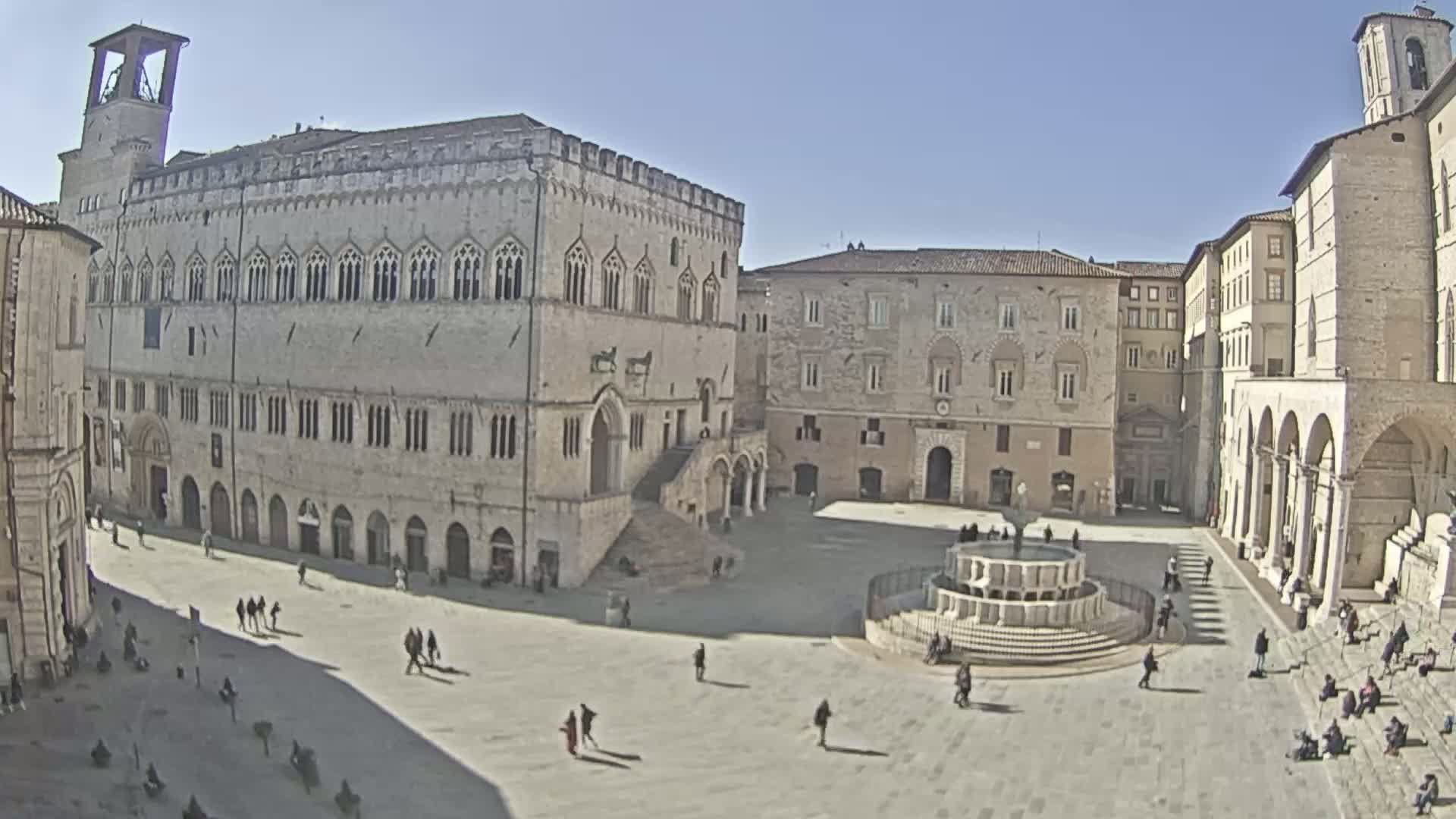 A large, mostly empty town square with several stone buildings, a fountain, and a few people scattered throughout, under sunny skies.