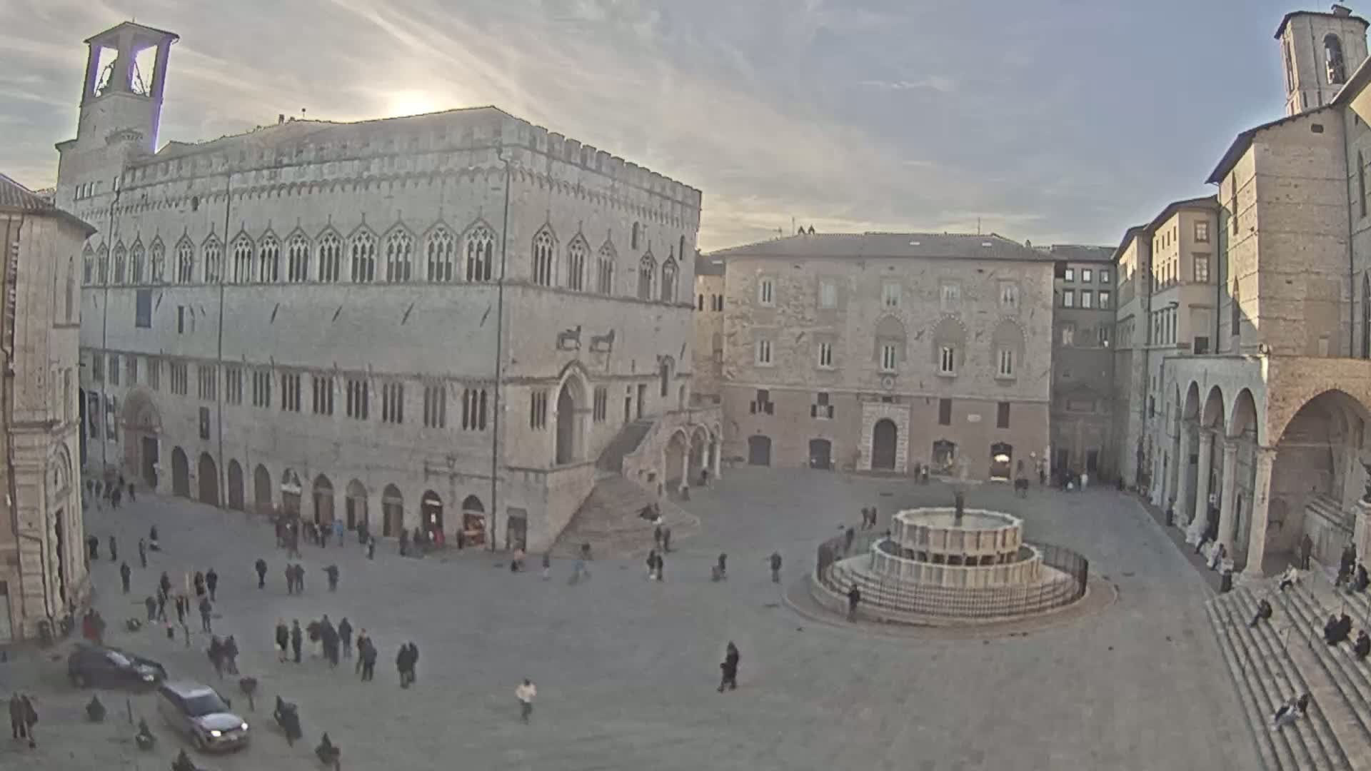 A large, stone public square with a fountain at its center is surrounded by historic buildings under a partly sunny sky.