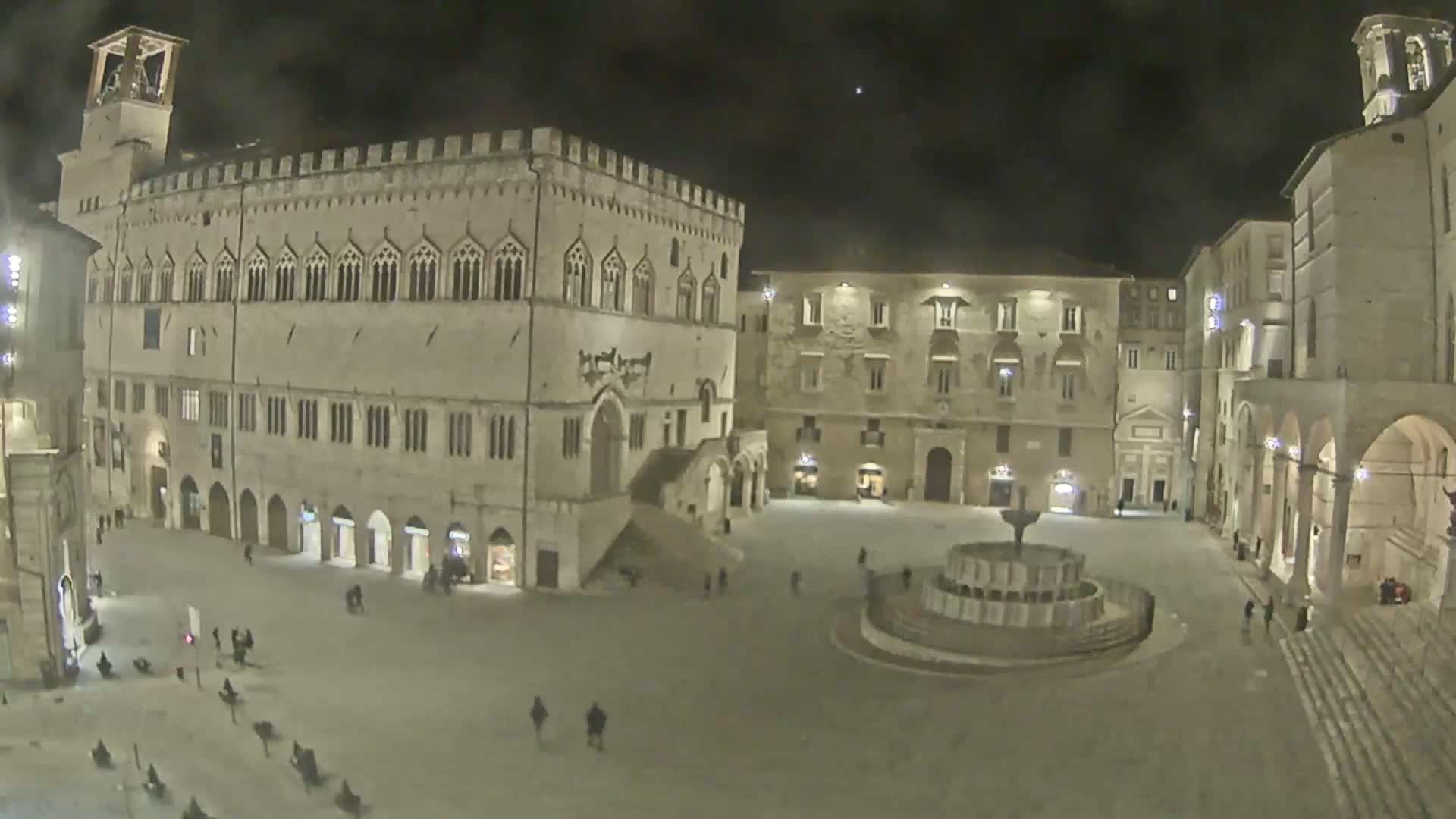 A large, mostly empty plaza at night, surrounded by historic stone buildings, features a central fountain.