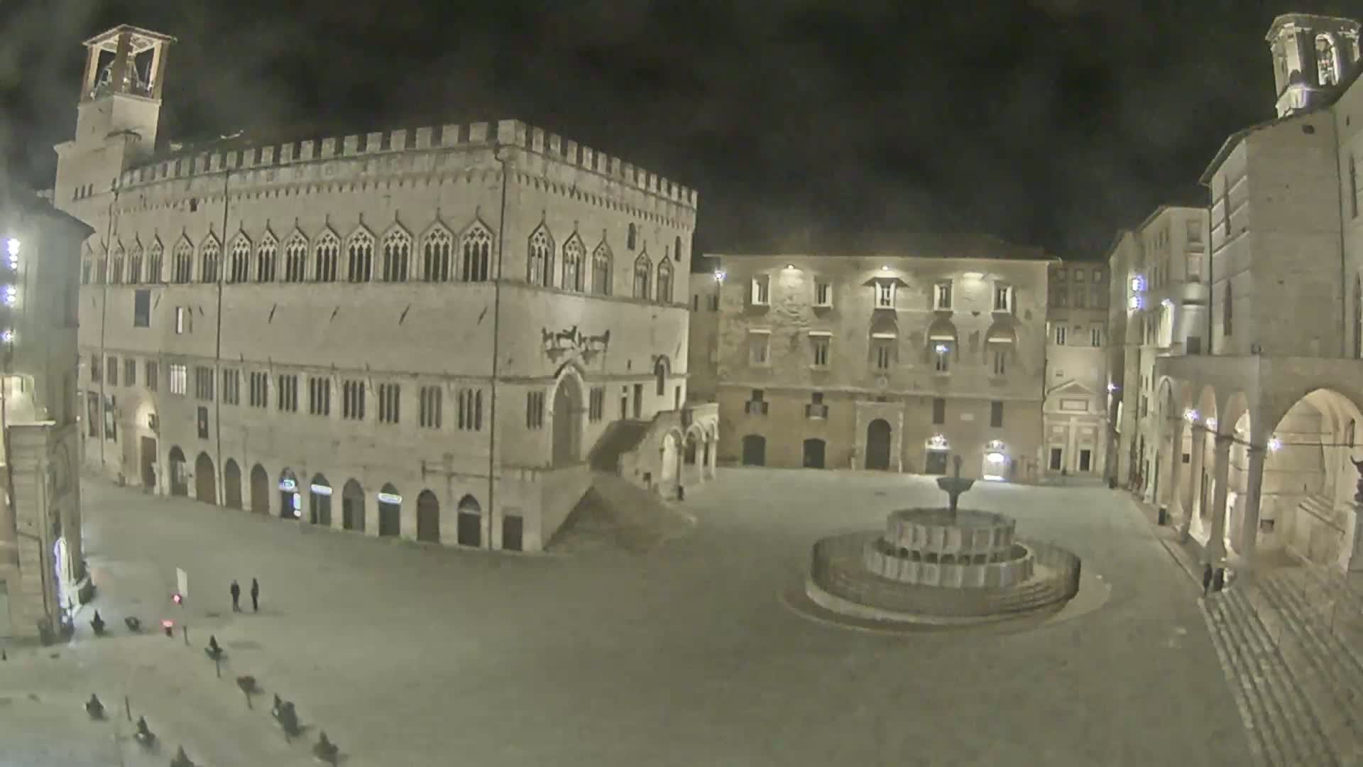 A large, empty town square at night, featuring a stone palazzo, other buildings, and a fountain, under a dark sky.