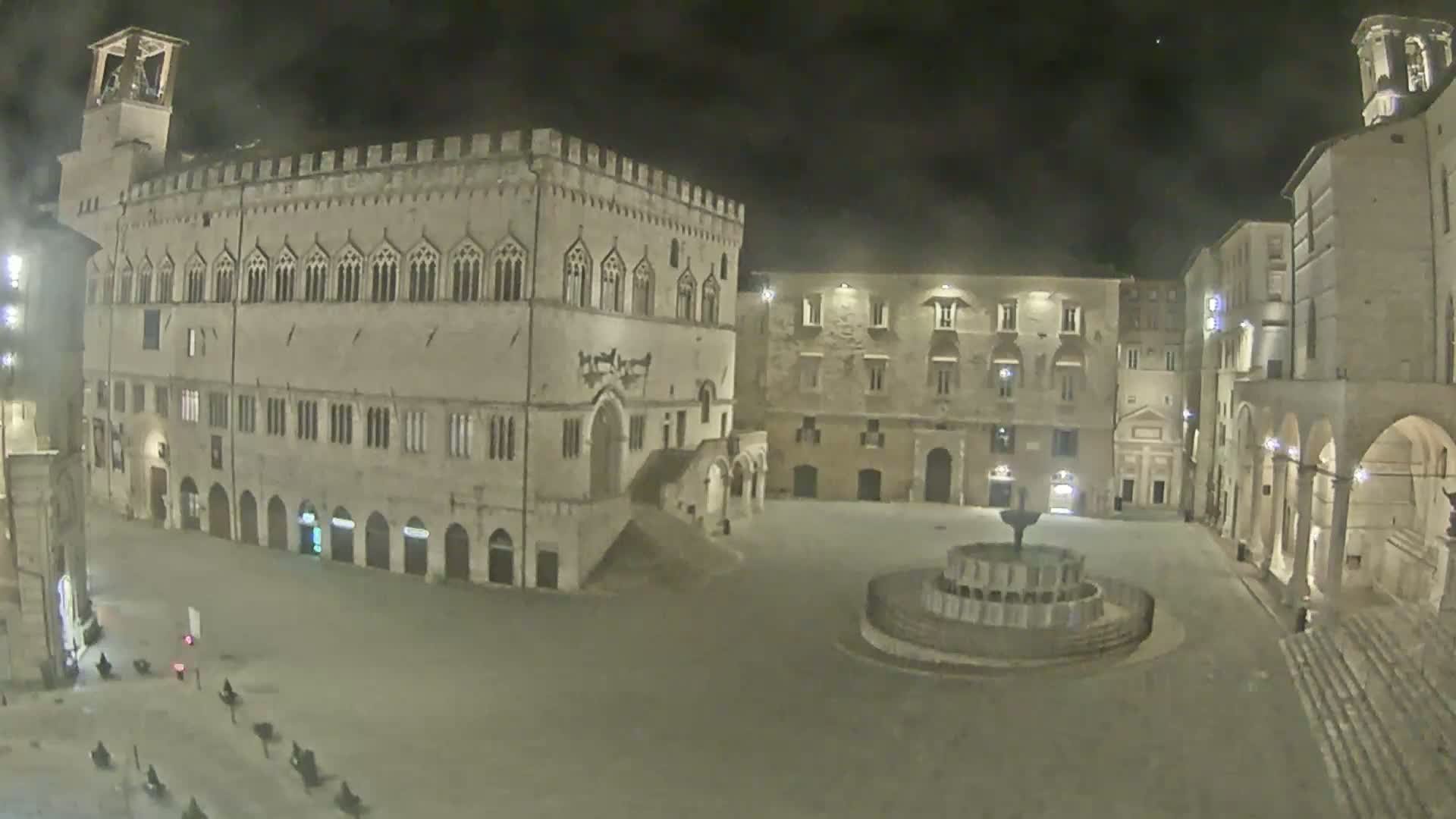 A large, empty town square at night, featuring a fountain and several illuminated stone buildings, one of which is a large palazzo.