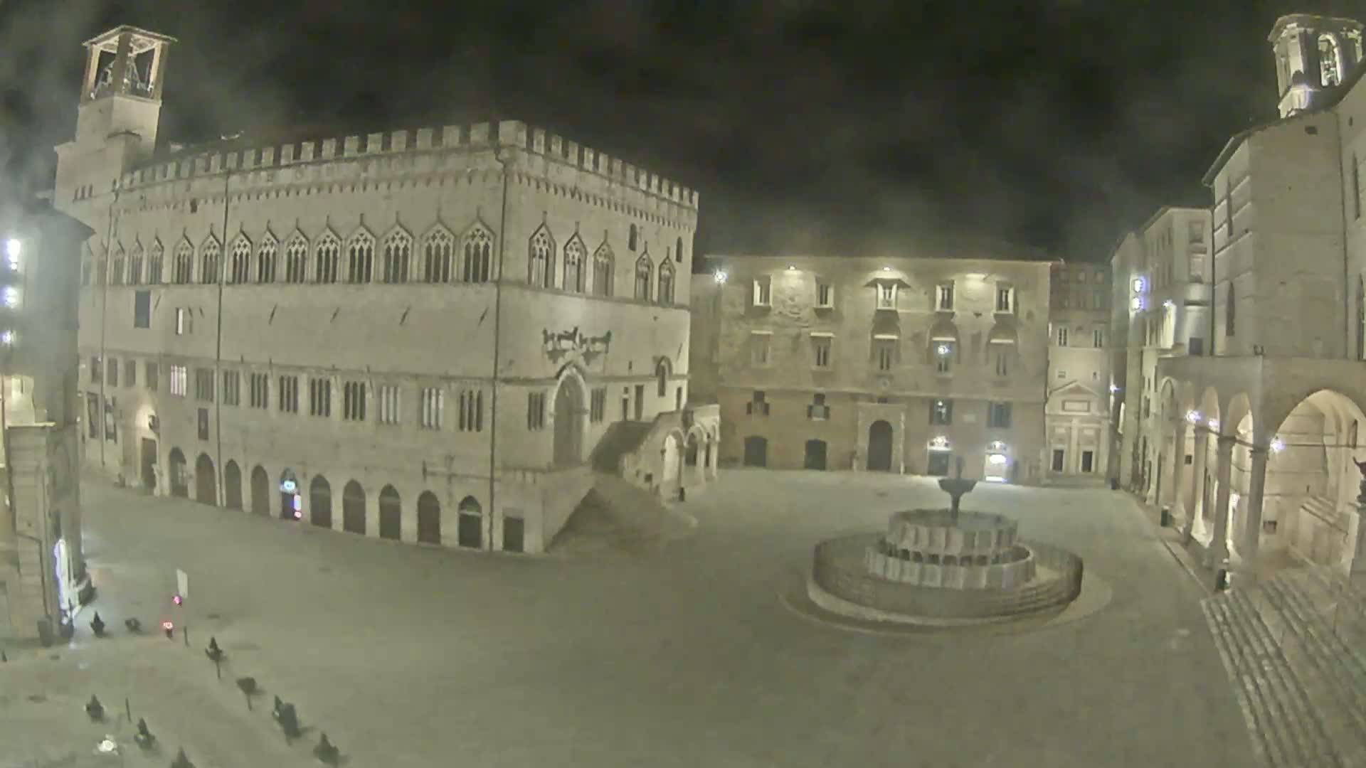 A large, empty town square at night, featuring a central fountain and several historic stone buildings, is illuminated by artificial light.