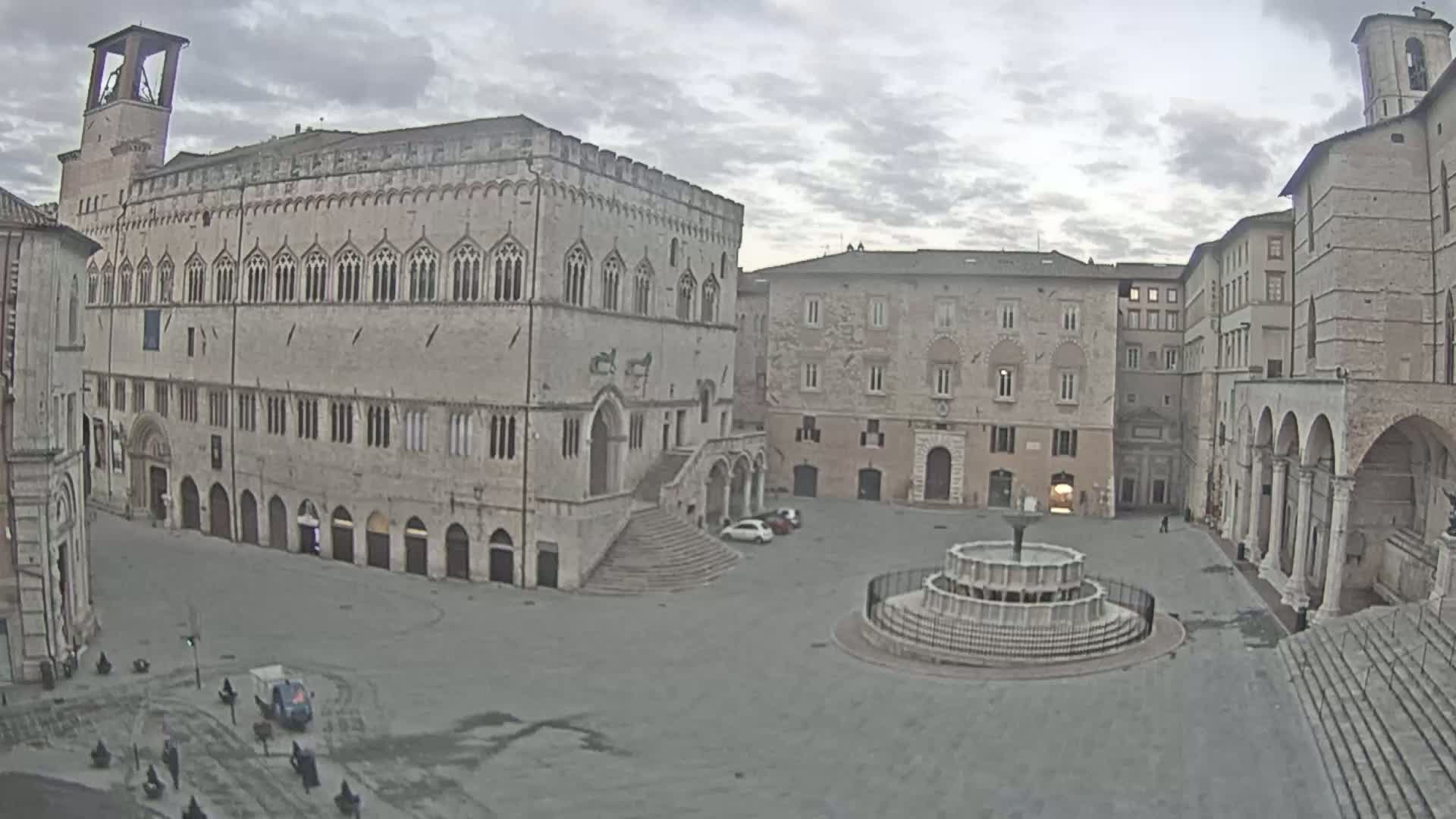 A large, mostly empty town square is surrounded by aged stone buildings under an overcast sky, featuring a circular fountain at its center.