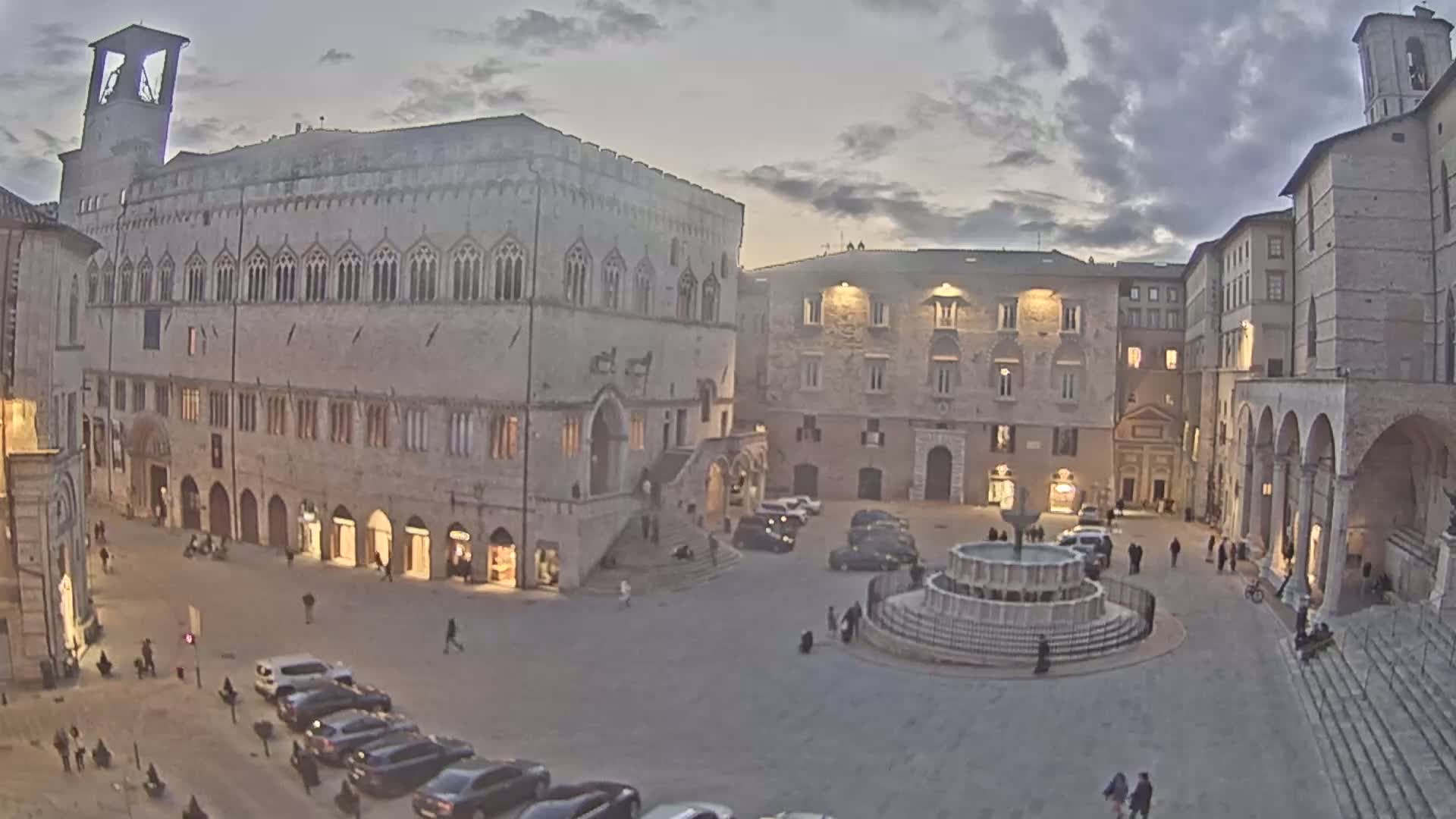 A town square, under a cloudy twilight sky, features a central fountain, surrounded by several stone buildings, and parked cars.