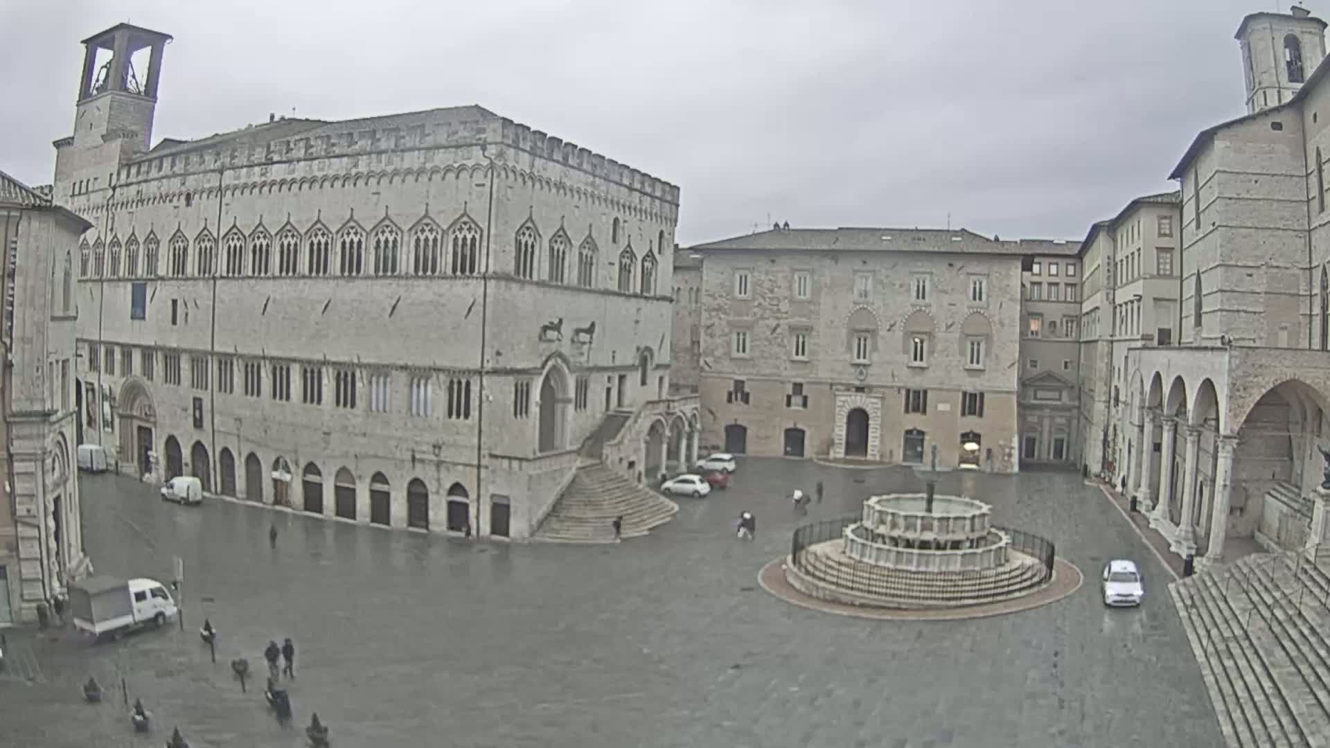 A large, stone plaza with a fountain at its center, surrounded by several historic buildings under an overcast sky.