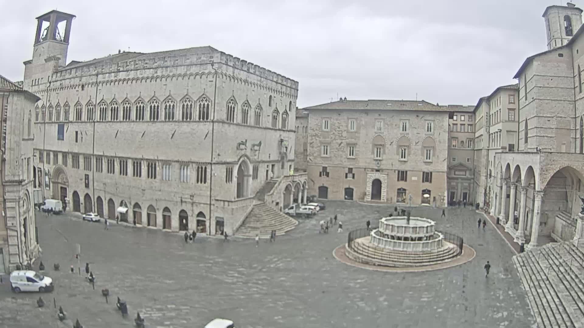 A large, stone plaza with a fountain at its center is surrounded by several historic buildings, under an overcast sky.