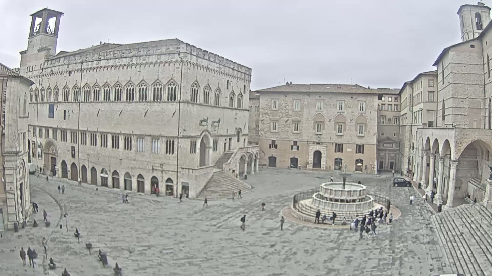 A large, mostly empty town square with several stone buildings, a central fountain, and a few people scattered throughout, under an overcast sky.