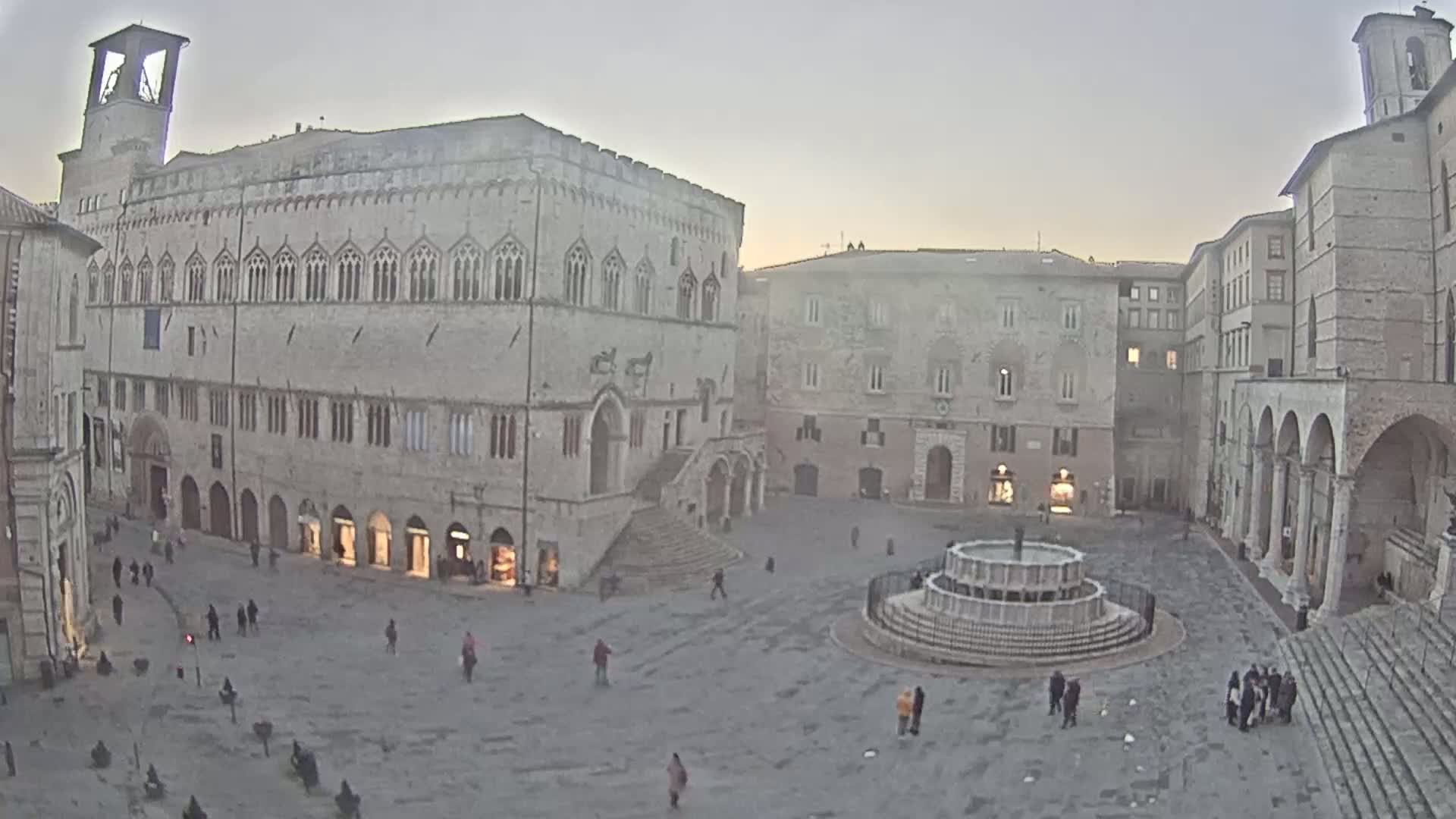 A large, stone plaza with a fountain at its center is surrounded by medieval-style buildings under a dusky sky.