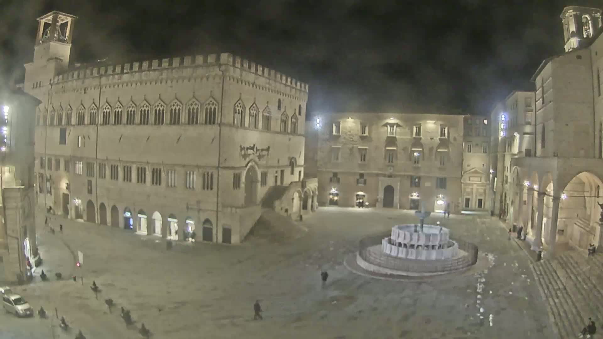 A nighttime view of a large, mostly empty town square surrounded by historic stone buildings, featuring a fountain in the center.