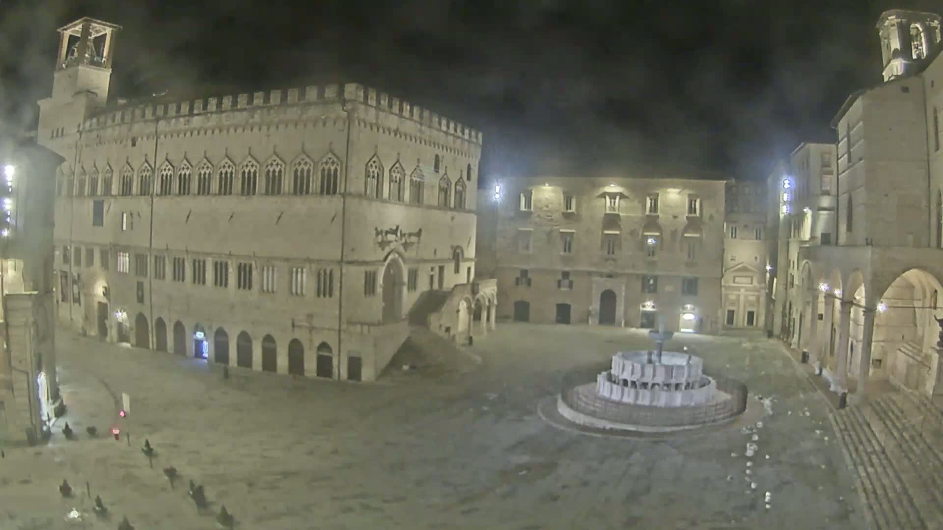 A mostly empty, stone plaza at night, featuring a large building and fountain, under a hazy sky.