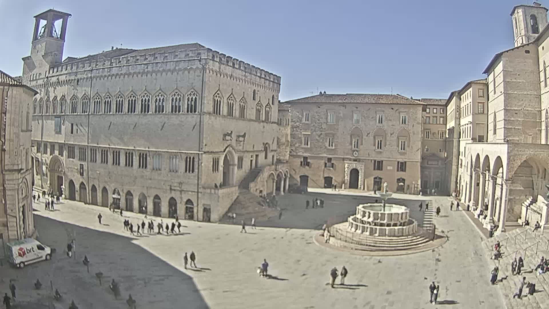 A large, sunlit town square features a central fountain and is surrounded by several stone buildings, one of which is a large palazzo.