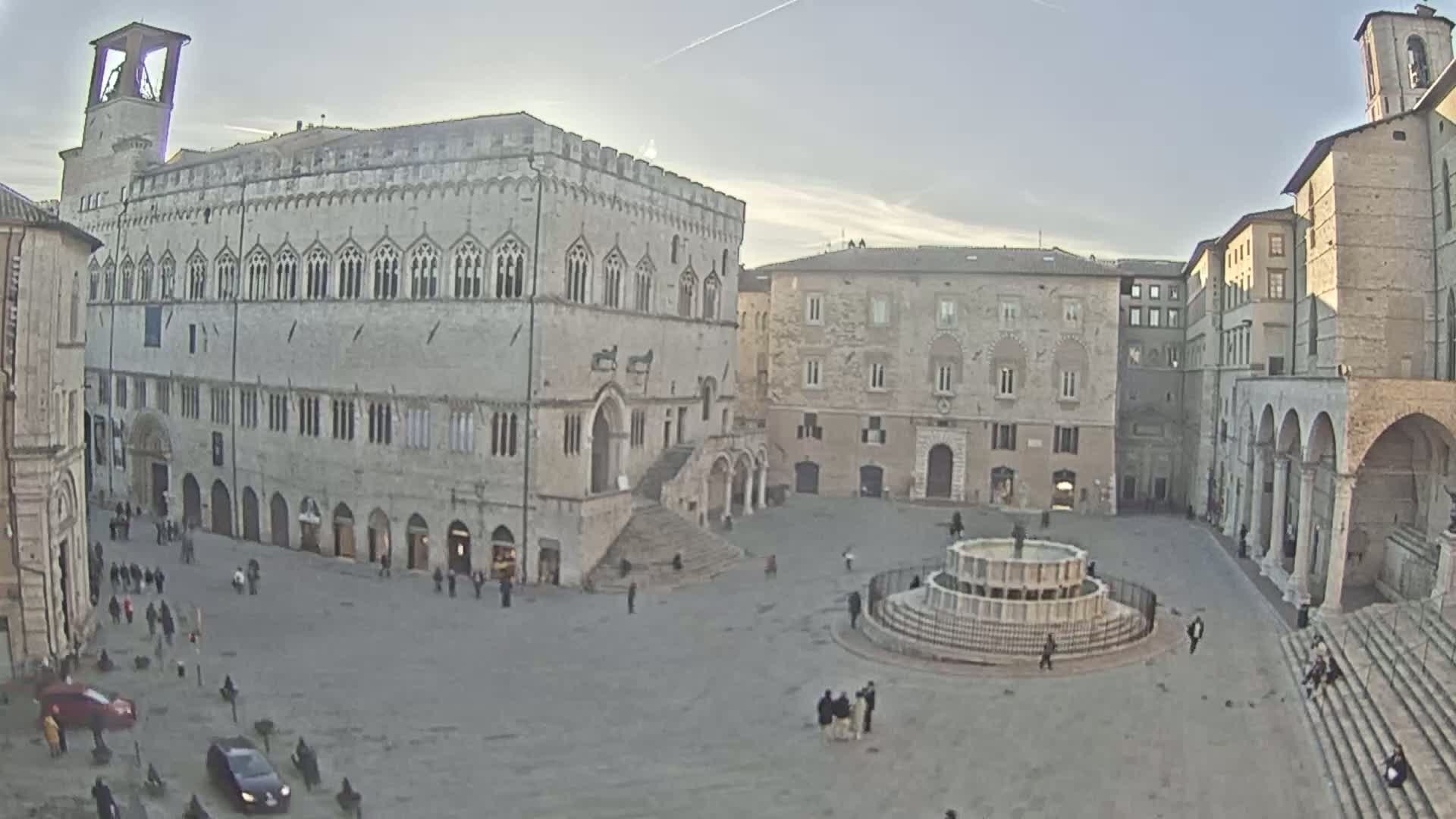 A large, mostly empty town square, under a mostly clear sky, is surrounded by several stone buildings, one of which has a prominent tower, and features a central fountain.