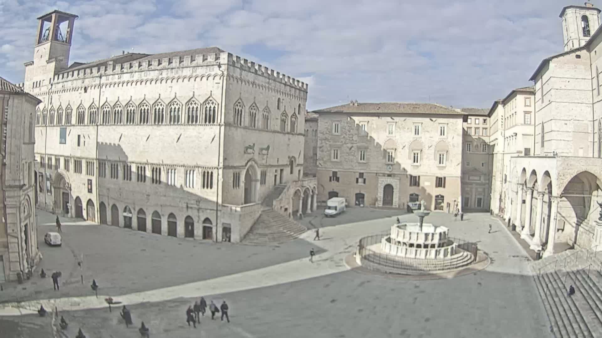 A large, mostly empty town square with a fountain and several stone buildings, under a mostly sunny sky.