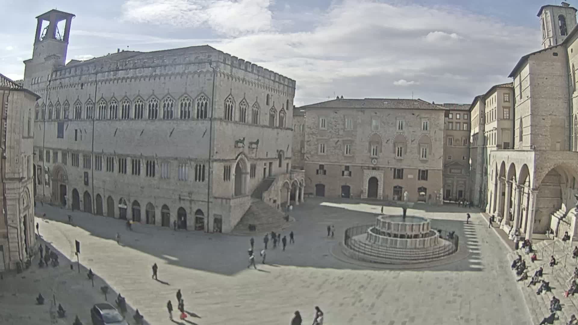 A large, mostly empty town square with a fountain in the center is surrounded by several historic stone buildings under a partly cloudy sky.