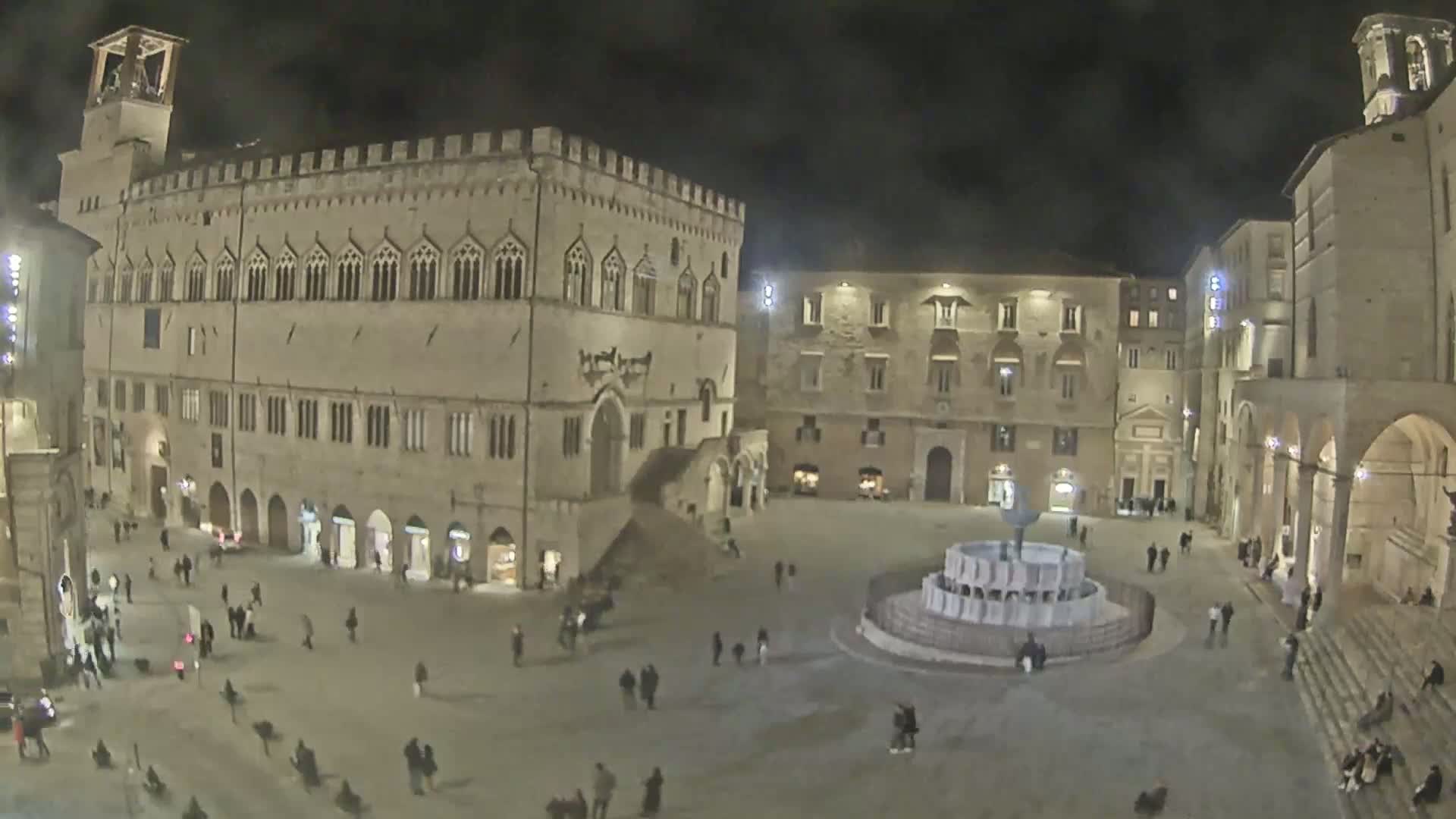A nighttime view of a large town square with a fountain at its center, surrounded by historic stone buildings, sparsely populated with people.