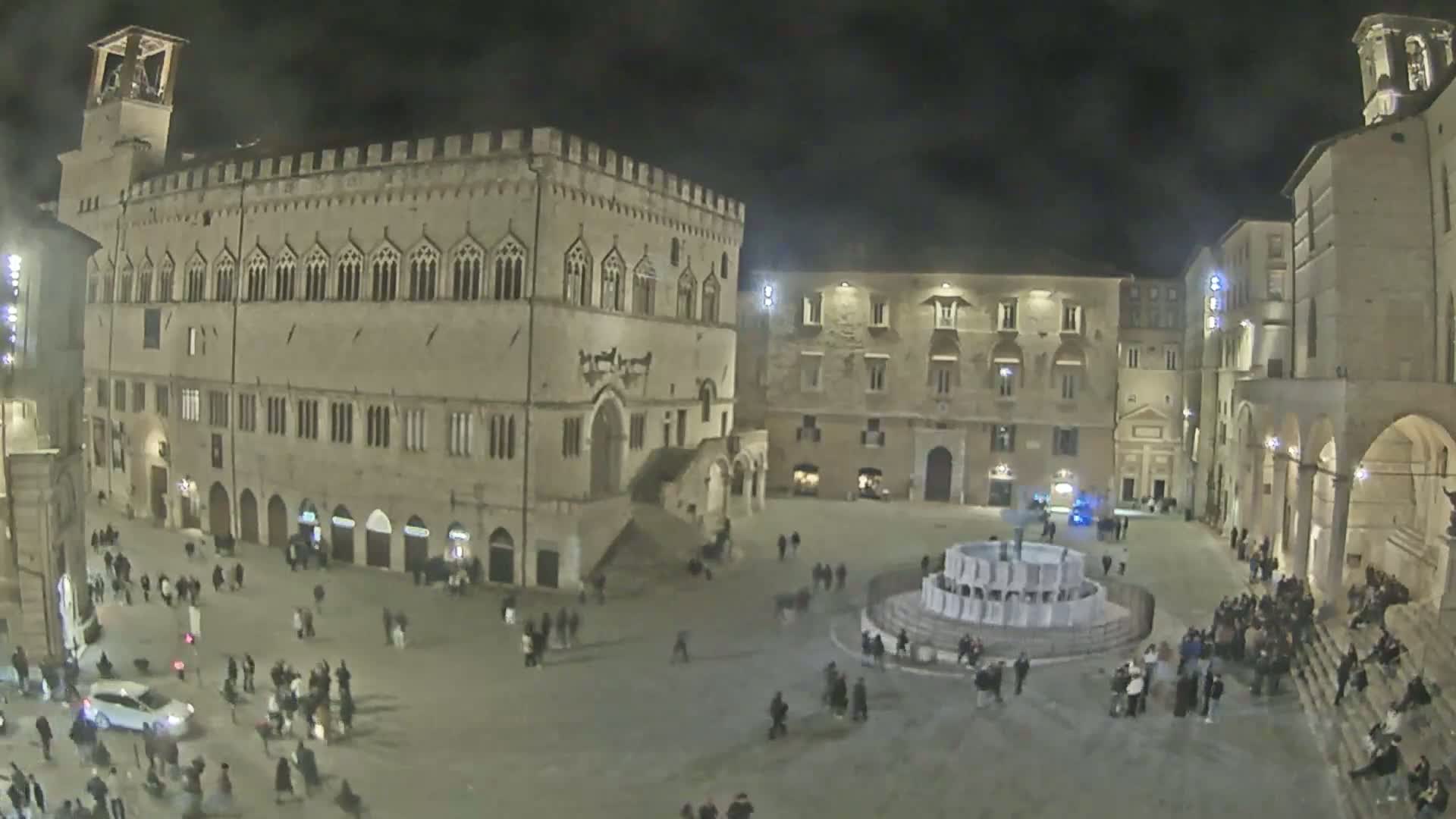 A nighttime aerial view shows a large town square with a fountain at its center, surrounded by several historical stone buildings and numerous people.