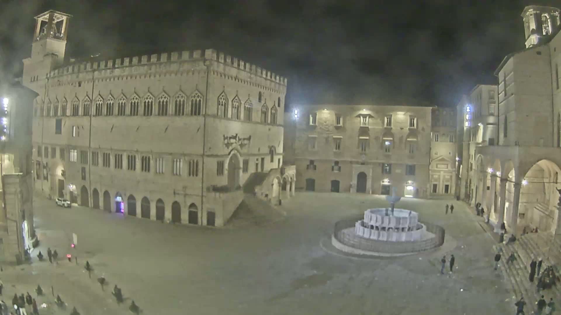 A nighttime view of a large, mostly empty plaza with a fountain in the center, surrounded by historic-looking buildings under a dark, cloudy sky.