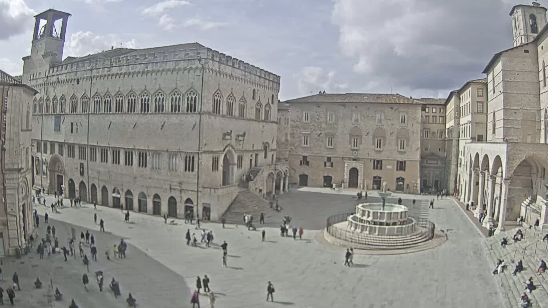 A large, light-stone plaza with a fountain at its center, surrounded by several multi-story buildings under a partly cloudy sky.