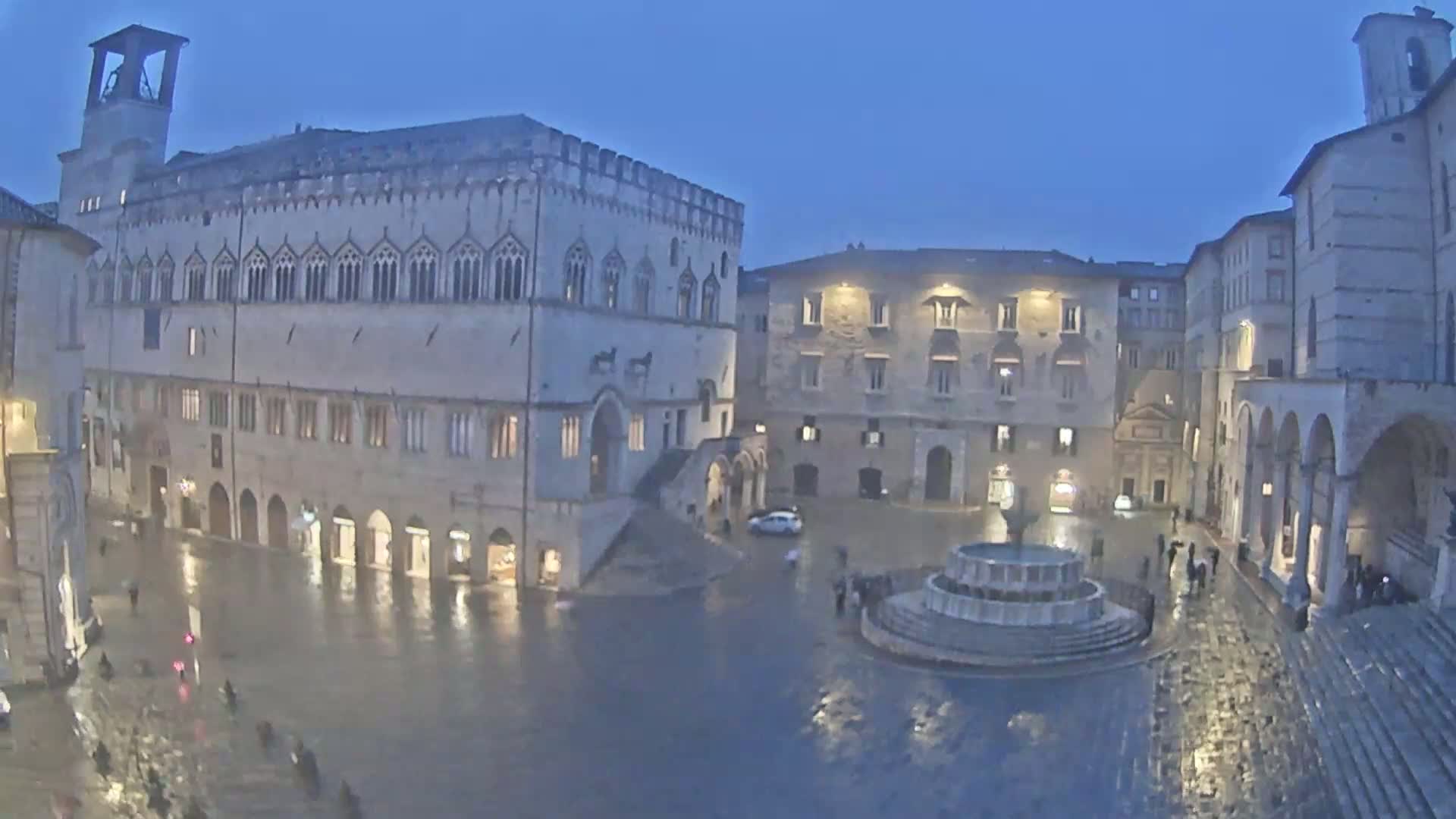 A rain-soaked town square at twilight, featuring a large stone building, a fountain, and several other buildings under dim lighting.