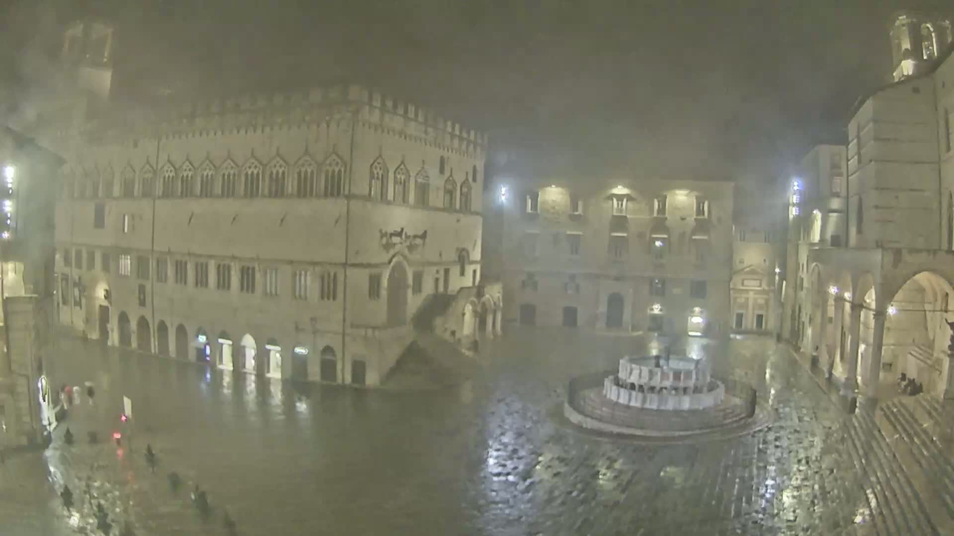 A large, flood-lit town square, partially submerged in rainwater at night, is surrounded by old stone buildings.