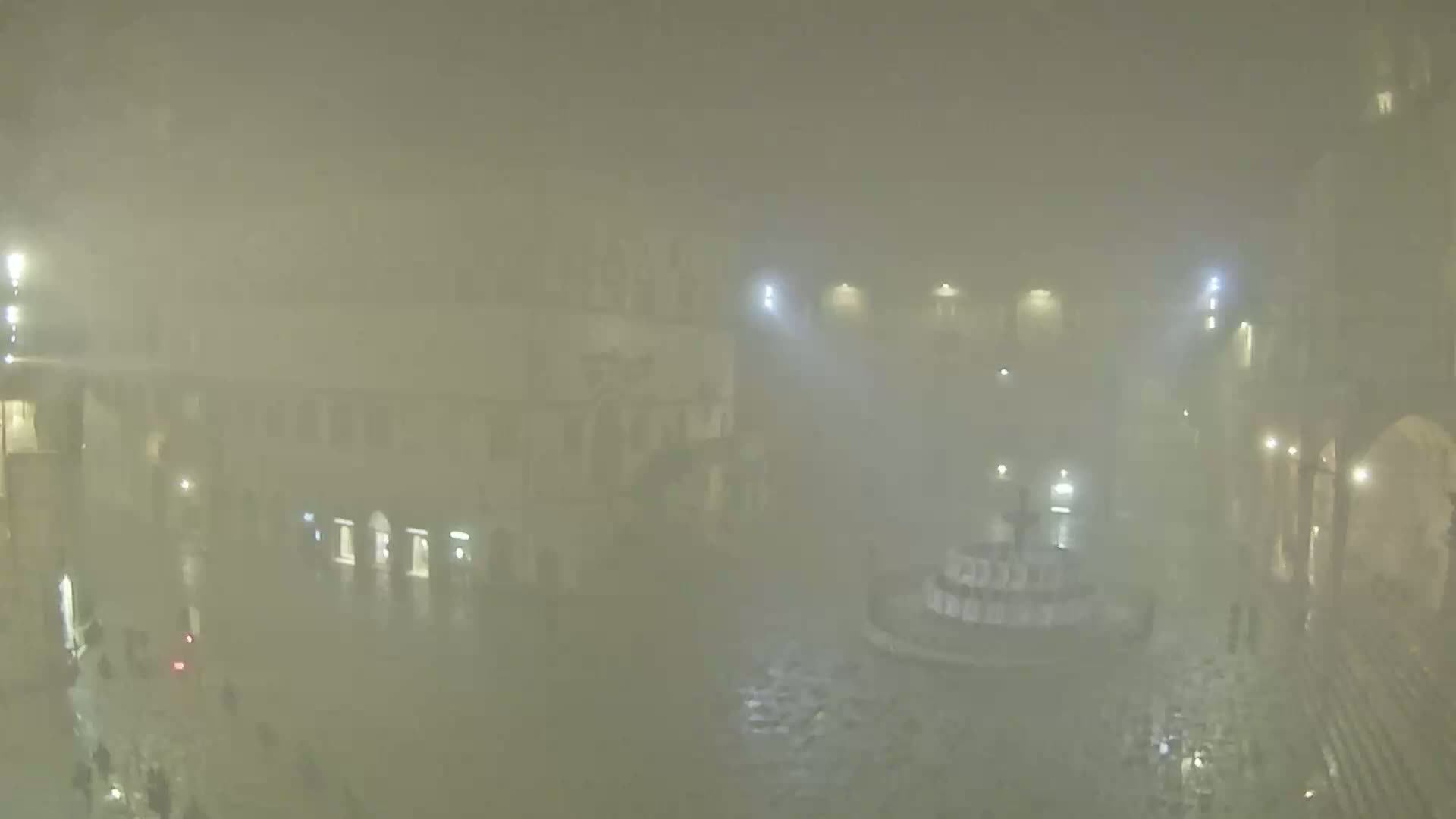 A nighttime, foggy view of a town square with a fountain in the center, surrounded by buildings and a few people.