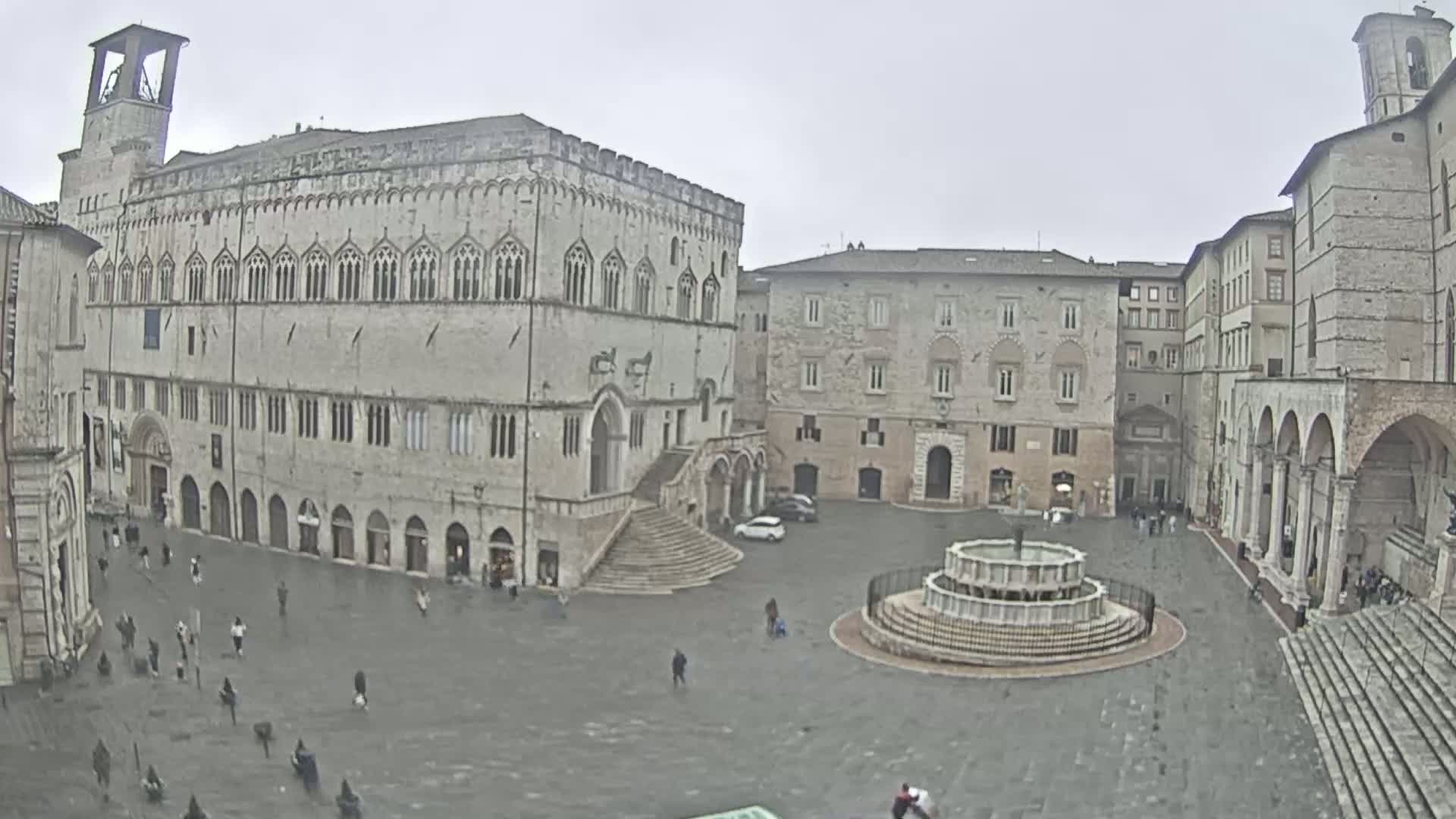 A large, mostly empty town square with several stone buildings, a central fountain, and a few scattered pedestrians on an overcast day.