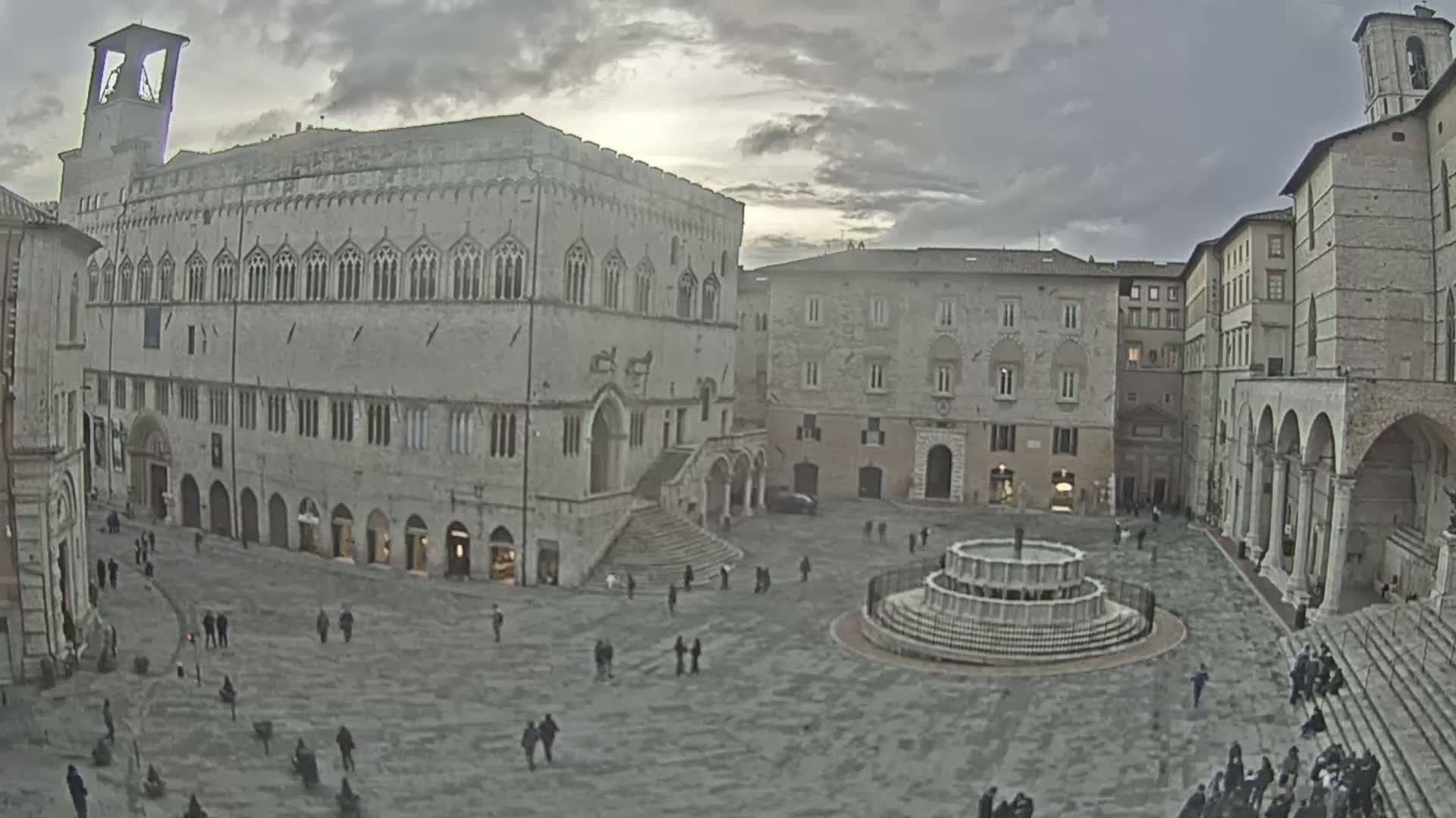 A large, stone plaza with a fountain at its center, surrounded by medieval-style buildings under a cloudy sky.