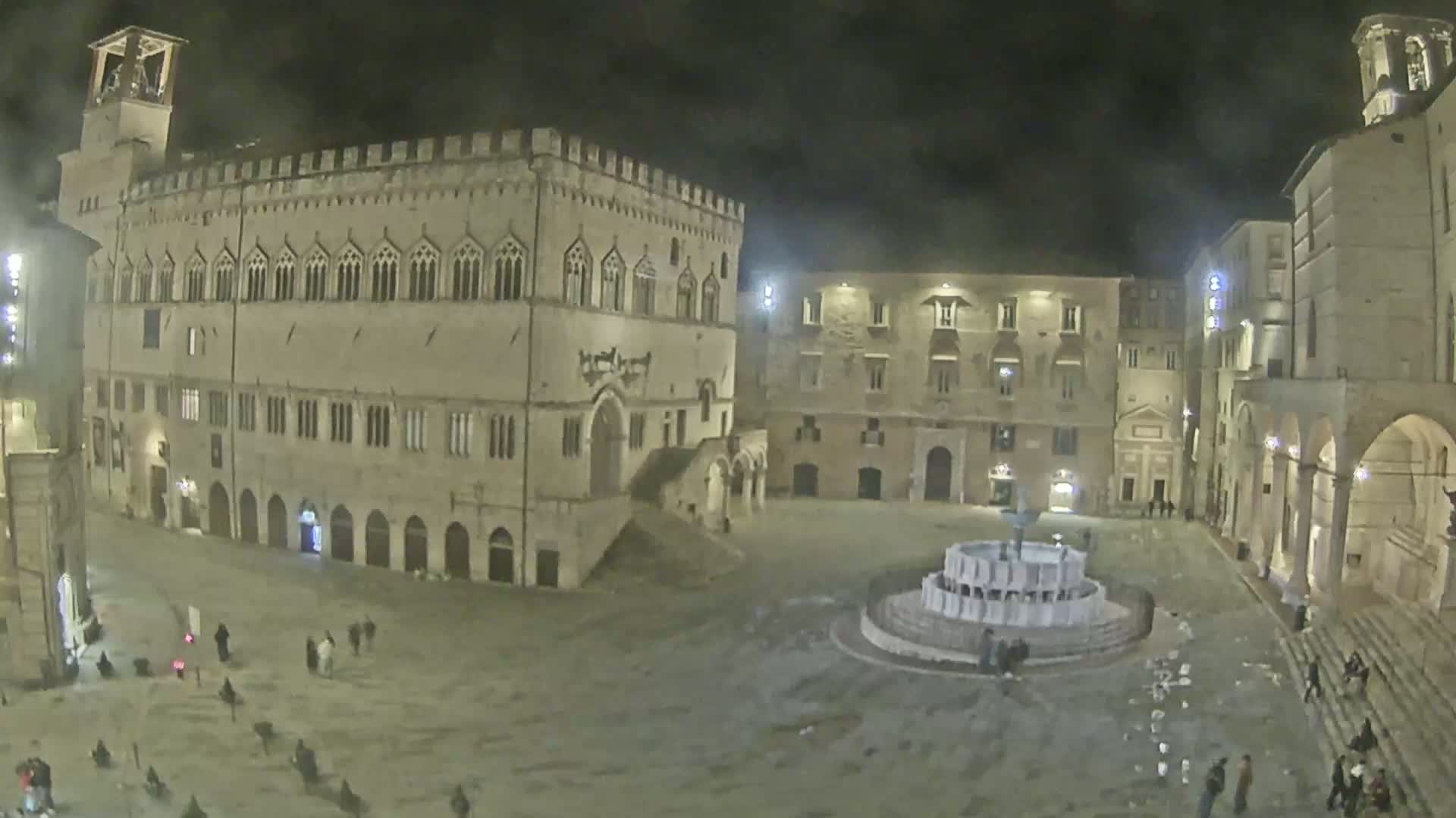 A nighttime view of a large, mostly empty town square with a fountain at its center, surrounded by historical-looking buildings.