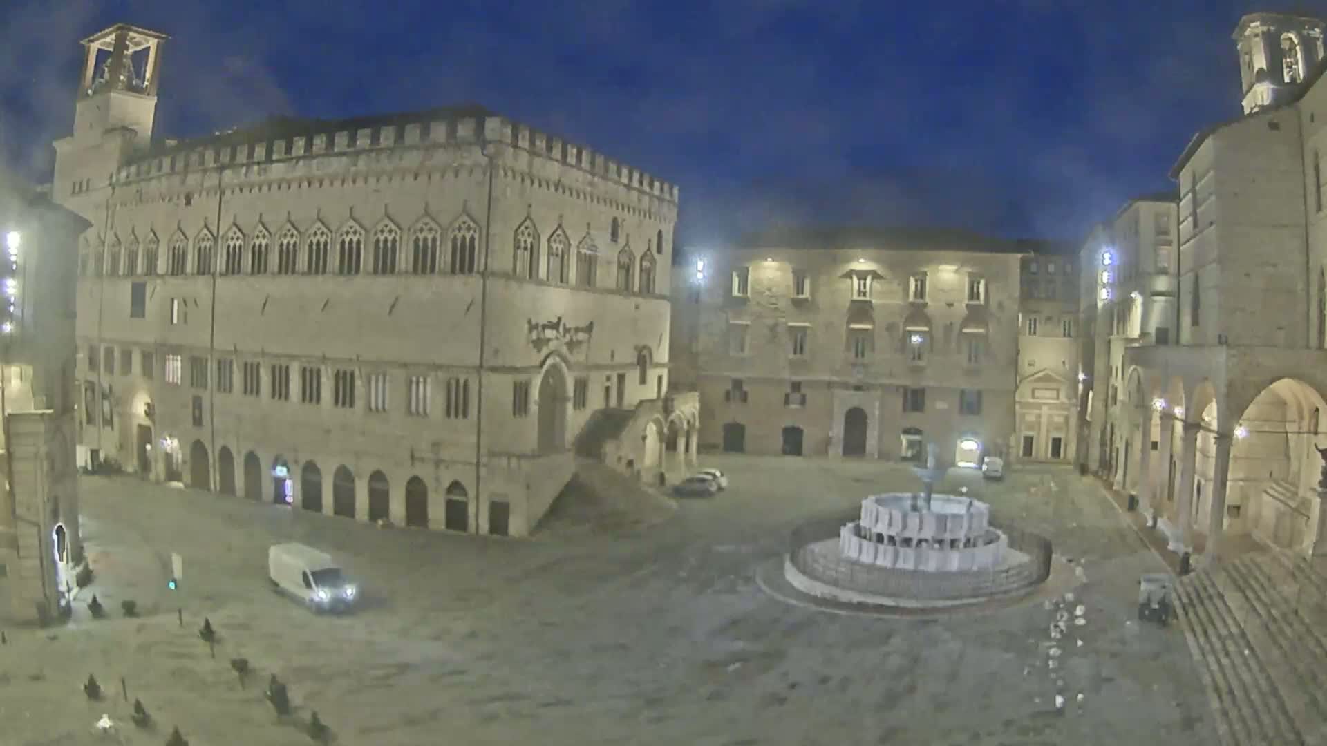 A large, empty town square at night, under a twilight sky, is surrounded by ancient stone buildings, one of which is a large palace, and features a fountain in the center.