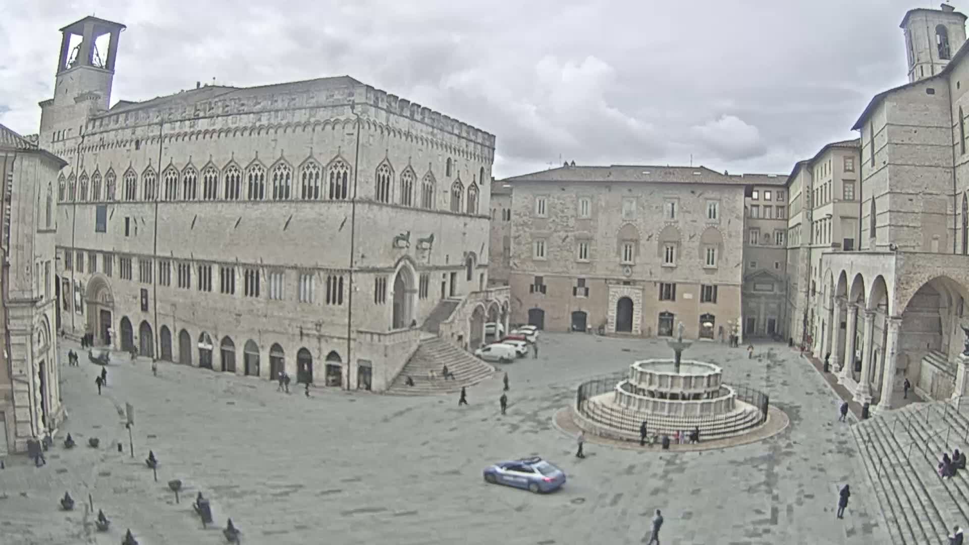 A large, mostly empty town square with a fountain at its center, surrounded by medieval-style buildings, under a cloudy sky.