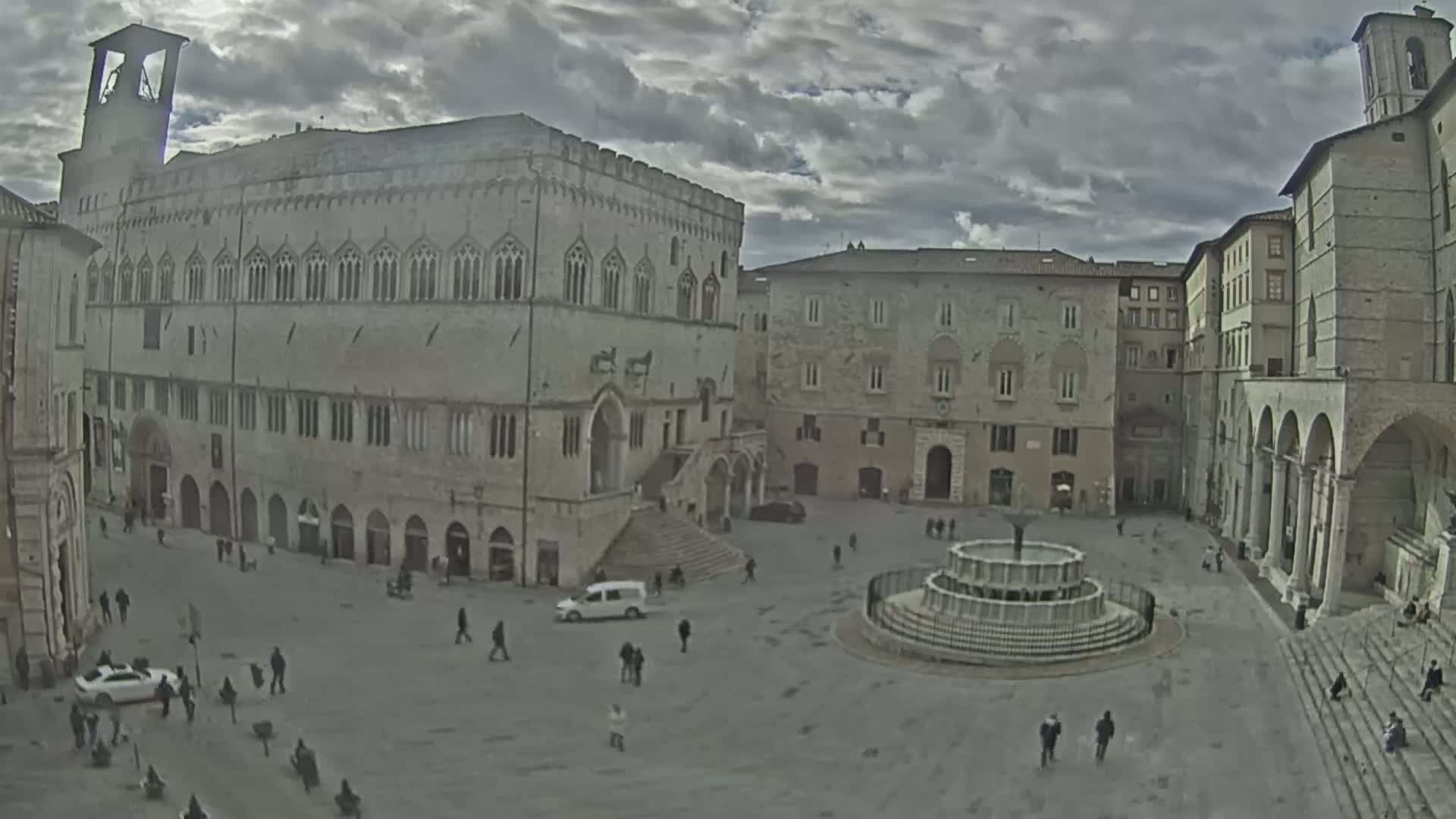 A large, mostly empty town square with a tiered fountain in the center is surrounded by several large stone buildings under a cloudy sky.