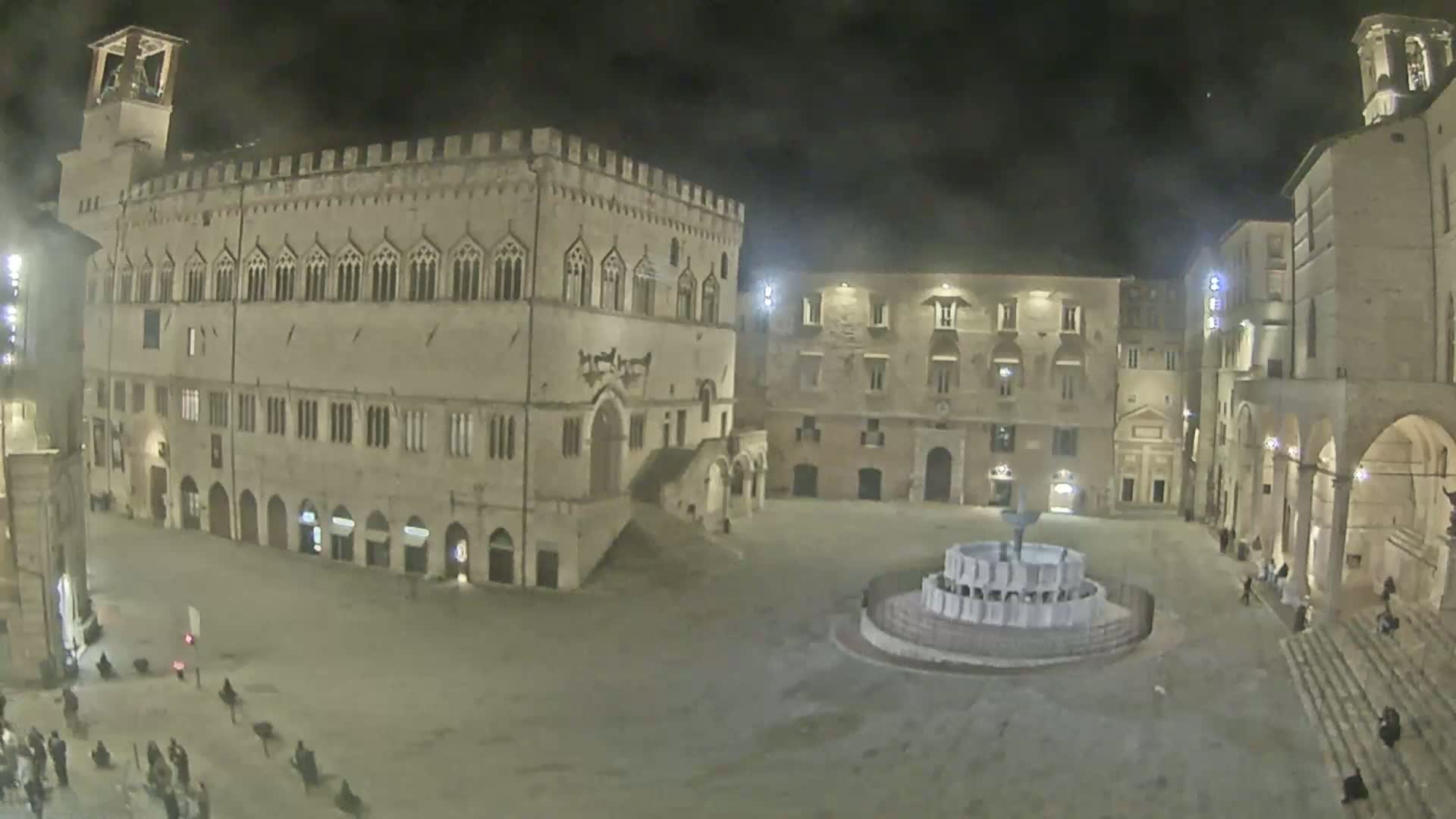 A nighttime view of a large, stone plaza with a fountain at its center, surrounded by historic buildings under a dark sky.