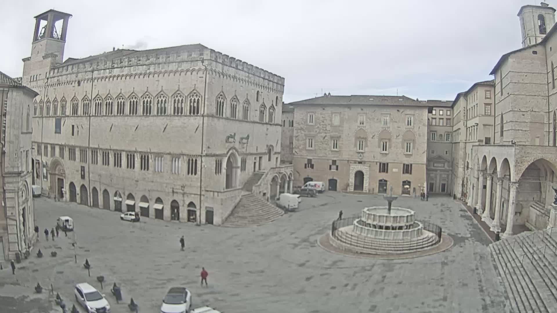 A large, stone piazza with a fountain in the center, surrounded by historic buildings, on an overcast day.