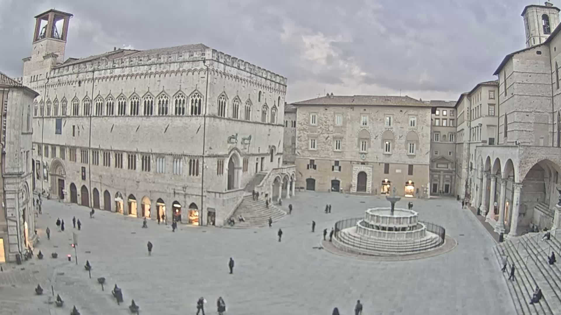 A large, mostly empty town square under an overcast sky features a central fountain and is surrounded by several large, light-colored stone buildings.