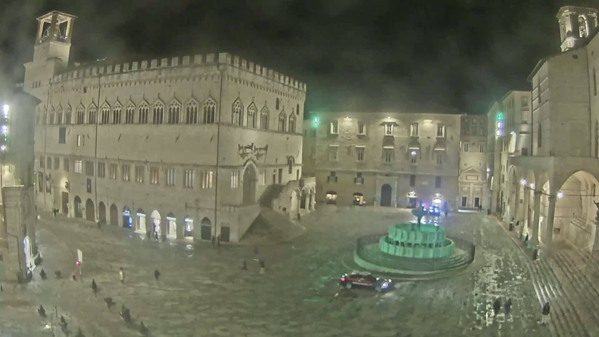 A large, illuminated town square at night in the rain, features a fountain and several historic-looking buildings.