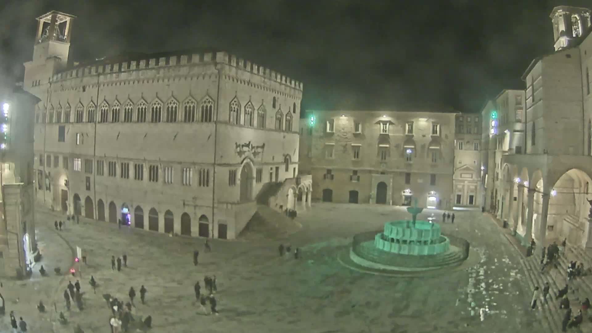 A large, sparsely populated town square at night, featuring a central fountain and several historic stone buildings under a dark sky.