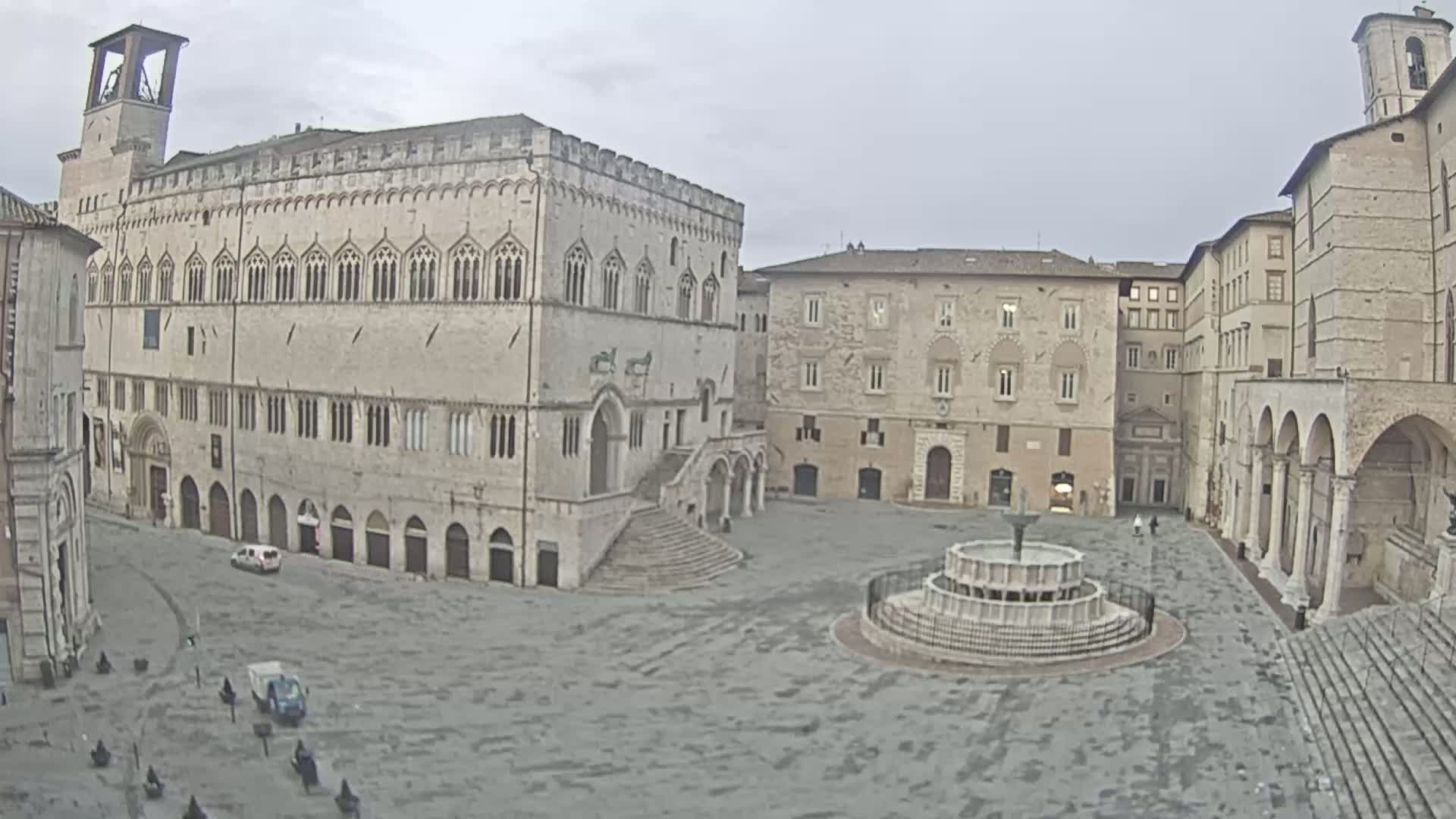 A large, mostly empty town square with several historic stone buildings, a fountain, and overcast skies.
