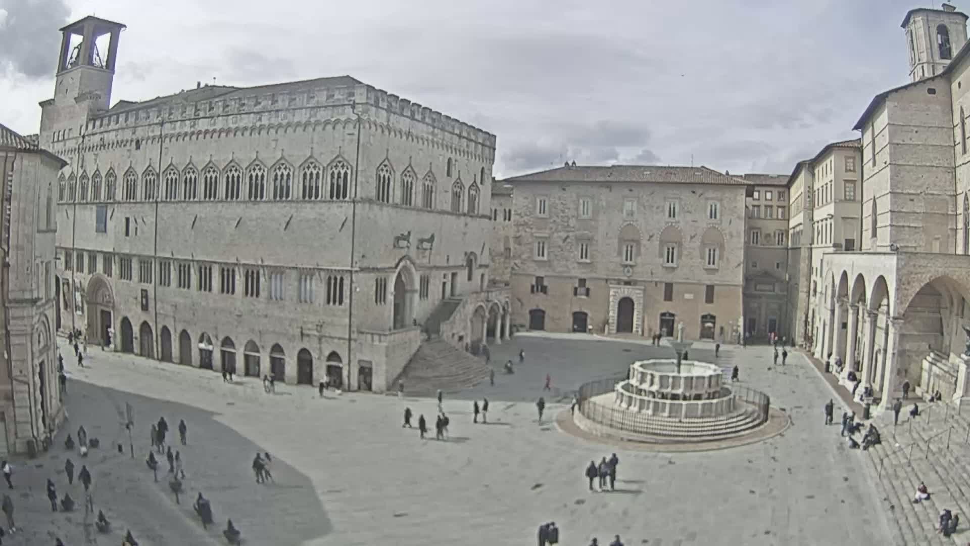 A large town square with several stone buildings surrounding it, including a large palace, under a mostly cloudy sky.