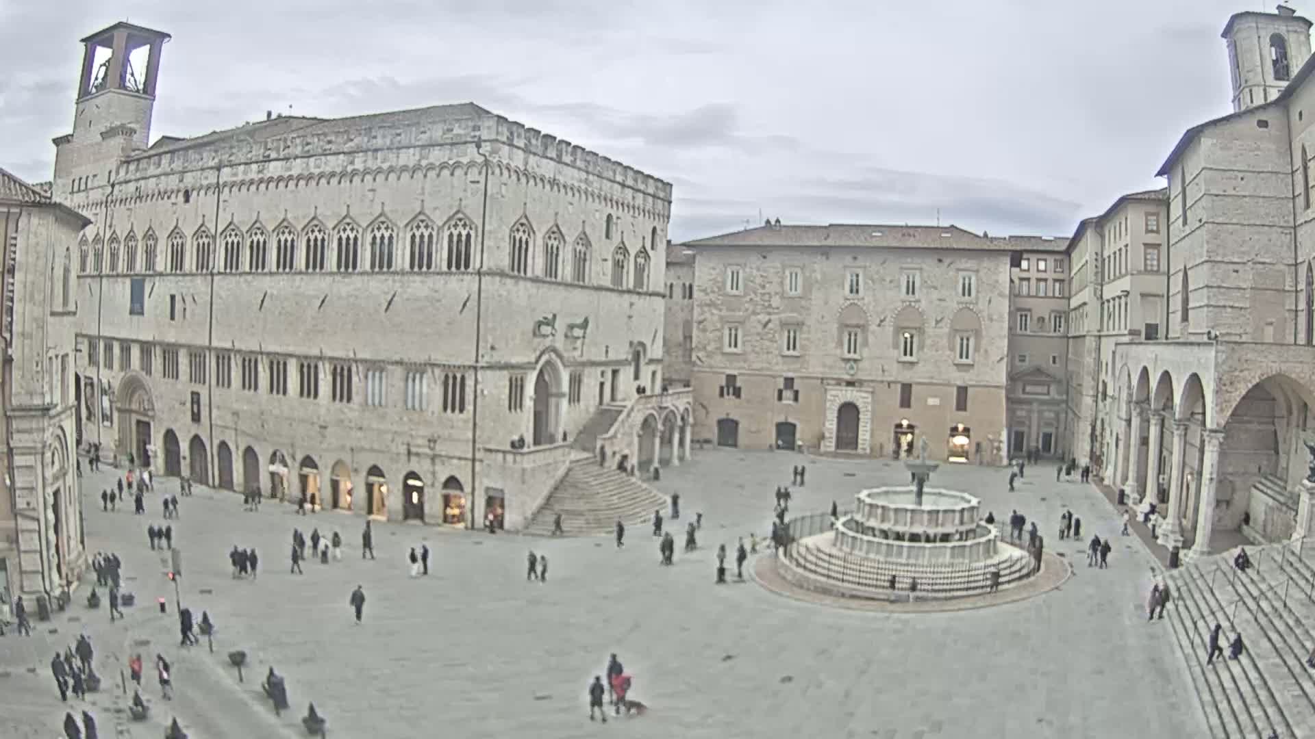 A large, mostly empty town square with several stone buildings, a central fountain, and a few people walking around on an overcast day.