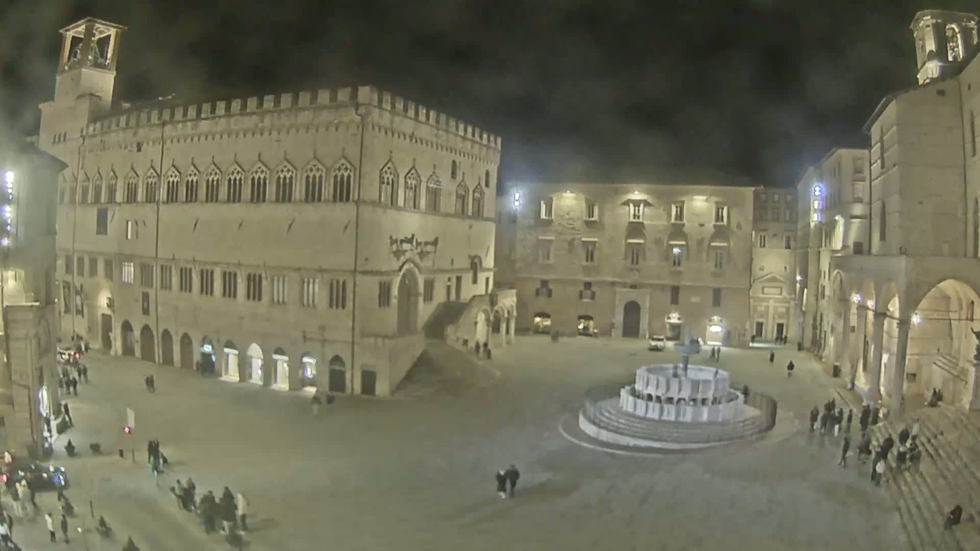 A nighttime view of a large town square, featuring several historic buildings, a fountain at the center, and a few people walking around.