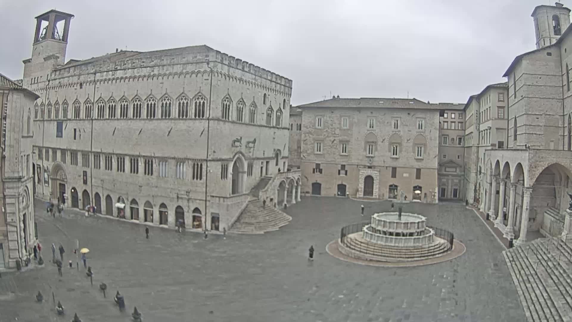 A large, mostly empty town square with stone buildings and a fountain, under an overcast sky.