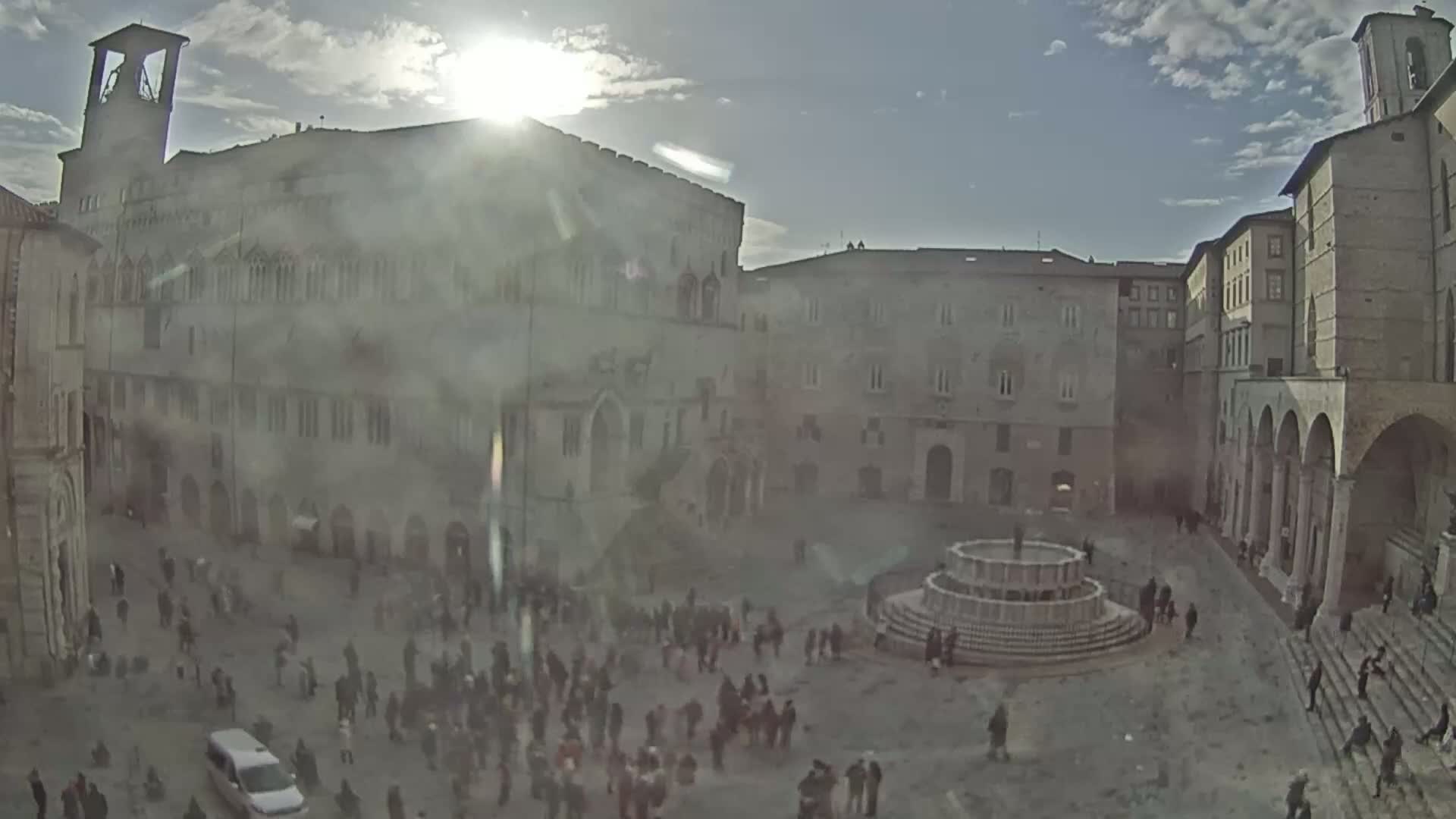 A sunny day reveals a large town square with a fountain at its center, surrounded by historical stone buildings and many people.