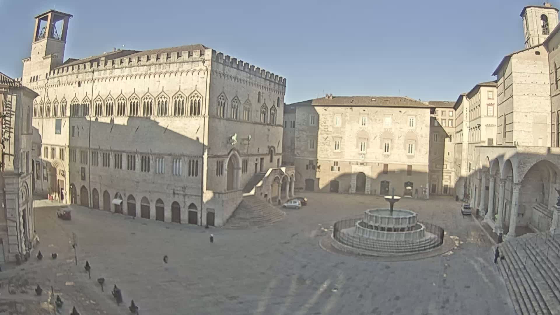 A large, empty plaza with a fountain is surrounded by several light-colored, historic buildings on a sunny day.