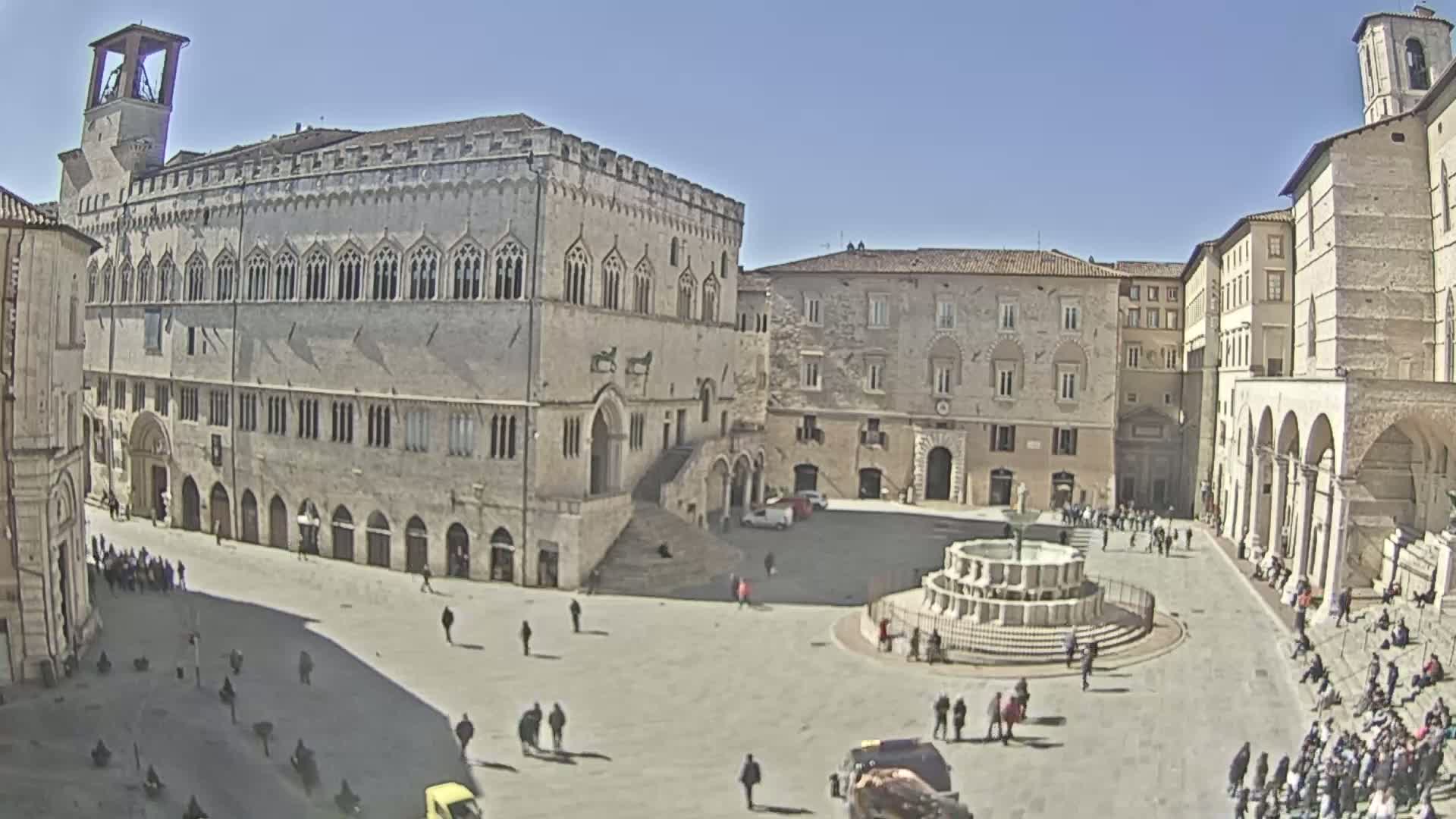 A large, sunny town square is surrounded by historic stone buildings, one of which is a grand palace with a staircase, and features a fountain in the center with several people walking and sitting around it.