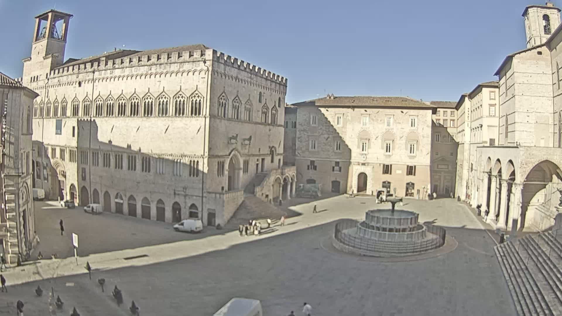 A large, sunny town square features a central fountain and is surrounded by several historic stone buildings, including a large palace.