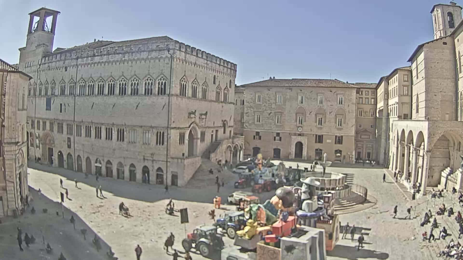 A large, sunlit town square with several historic buildings, a fountain, and a display of tractors and colorful carnival-like structures.