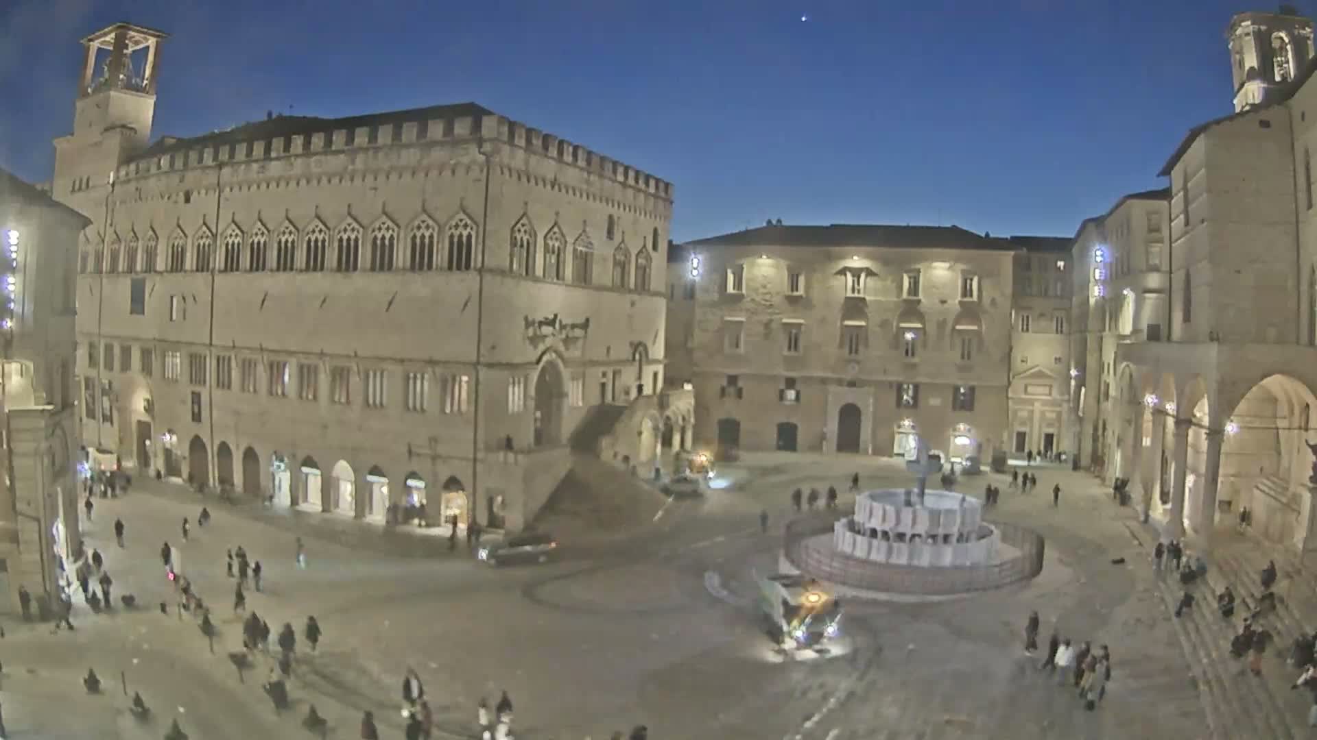 A large, stone plaza at night, featuring a central fountain and several historical buildings, is sparsely populated with pedestrians under a clear, dark sky.