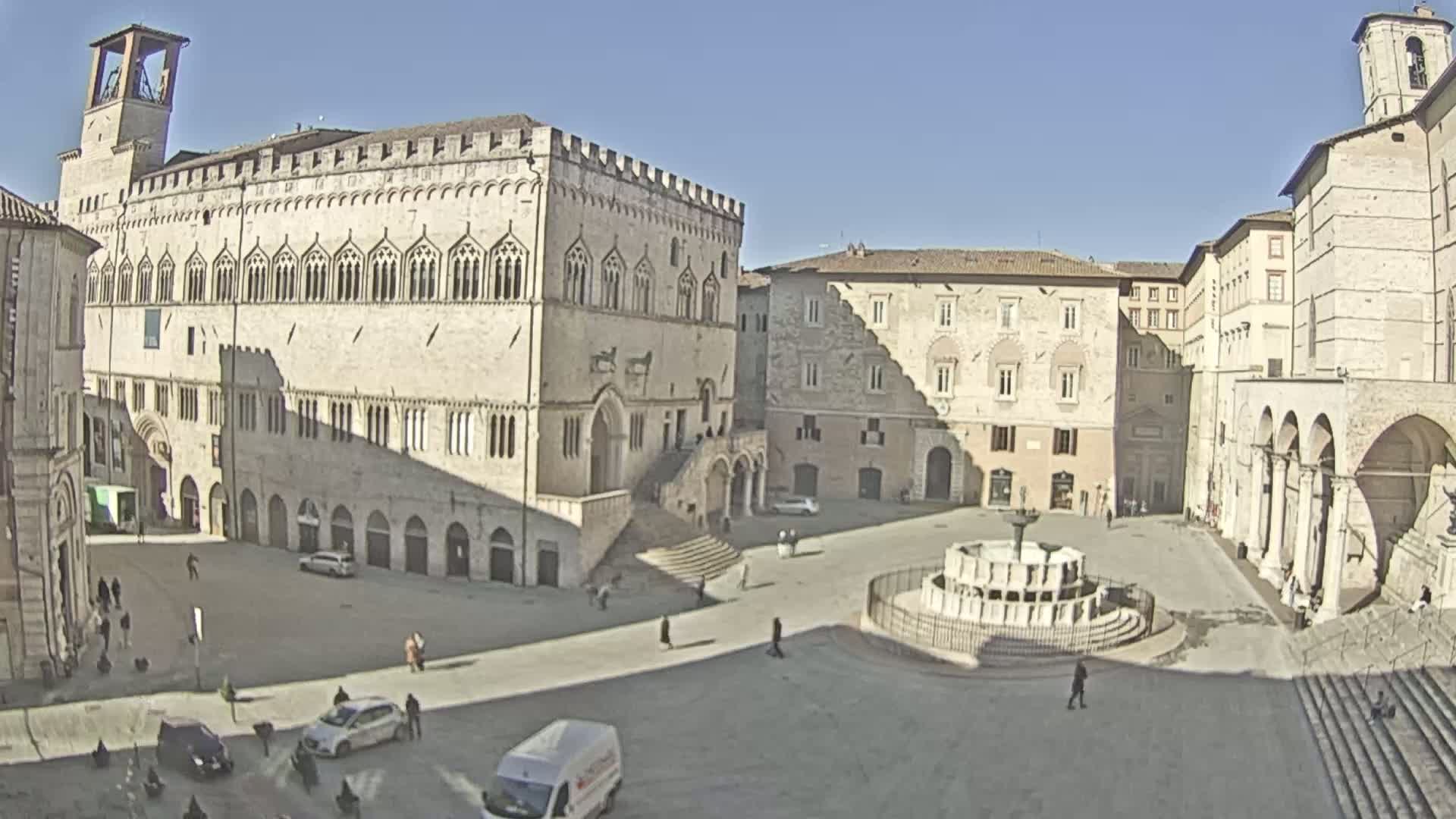 A sunny day reveals a large, open square surrounded by historical stone buildings, one prominently featuring a grand staircase, and a central fountain.