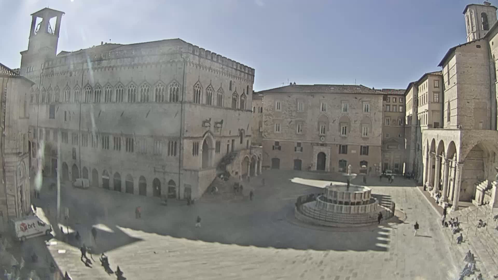 A sunny day reveals a large, stone town square with a fountain at its center, surrounded by historic buildings, including a large palace and church.