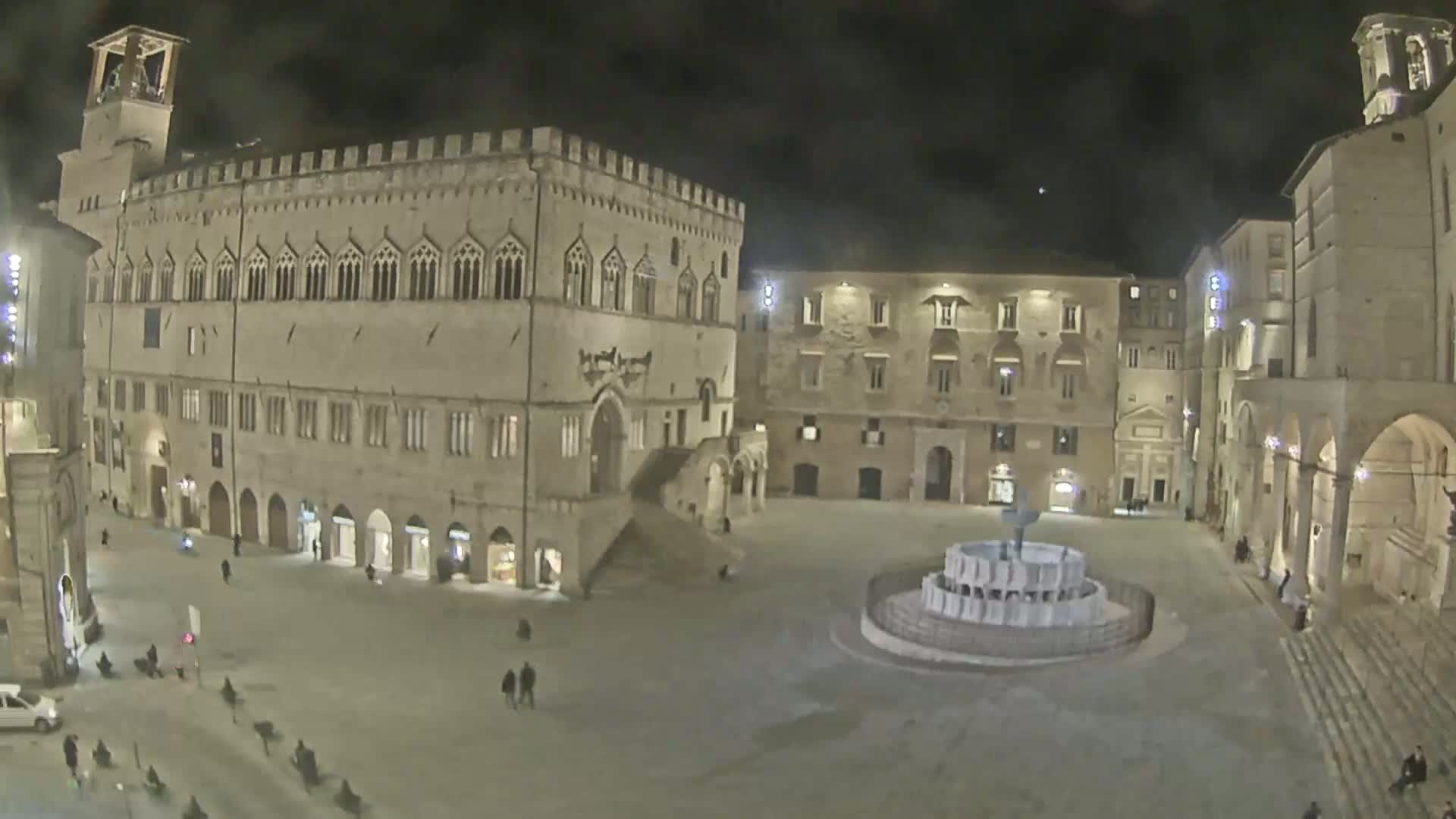 A nighttime view of a large, stone plaza with a fountain in the center, surrounded by historic buildings.